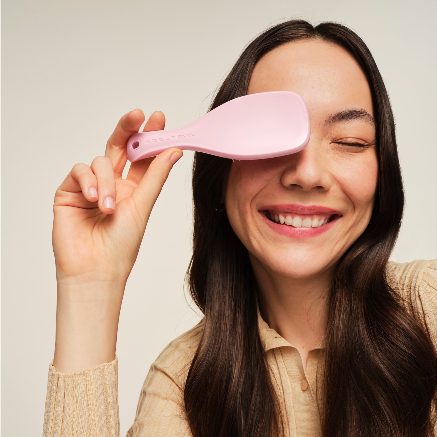 Eine lächelnde Frau hält eine rosafarbene Tangle Teezer Haarbürste vor ihr rechtes Auge, ihr langes, dunkles Haar fällt über ihre Schultern.