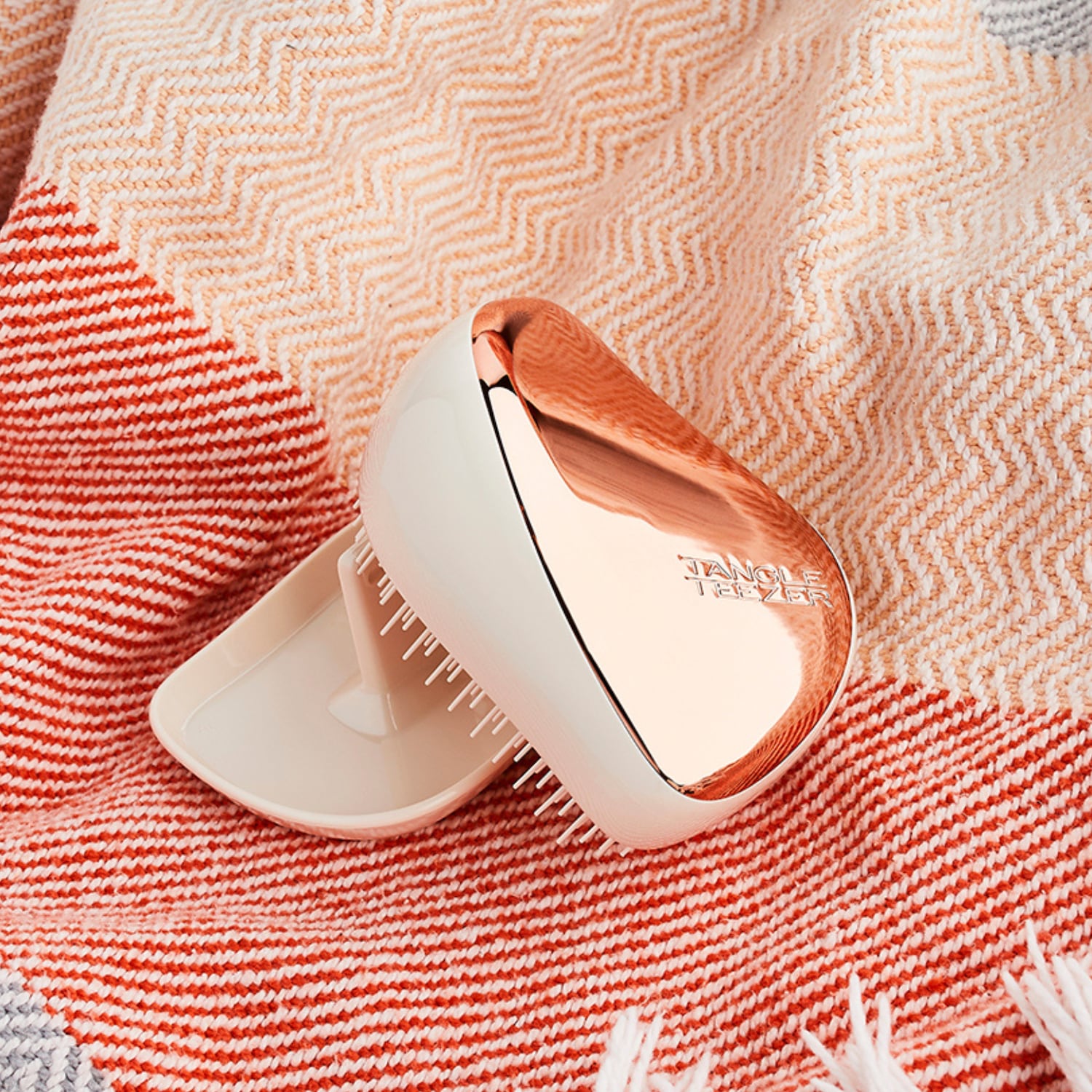 Der Tangle Teezer Compact Styler in Roségold und Creme liegt auf einem rot-weiß gestreiften Handtuch und ist bereit für müheloses Entwirren unterwegs.