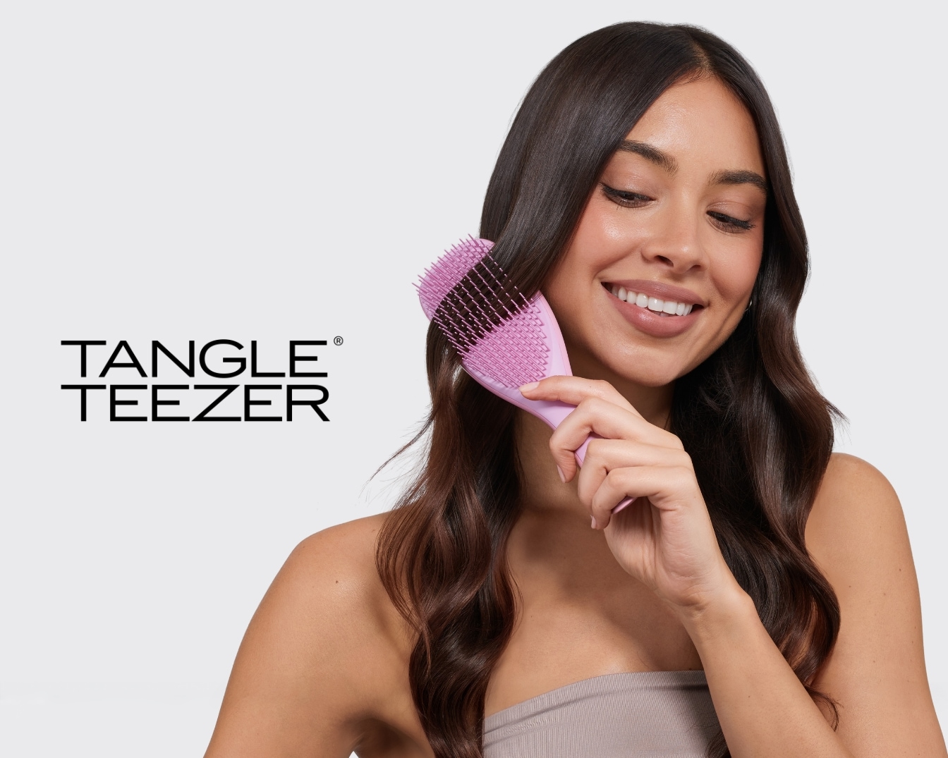 Eine lächelnde Frau mit langen, dunklen Haaren kämmt sich mit einer lila Tangle Teezer Bürste, die sanft durch ihr Haar gleitet, während das Tangle Teezer Logo links im Bild zu sehen ist.