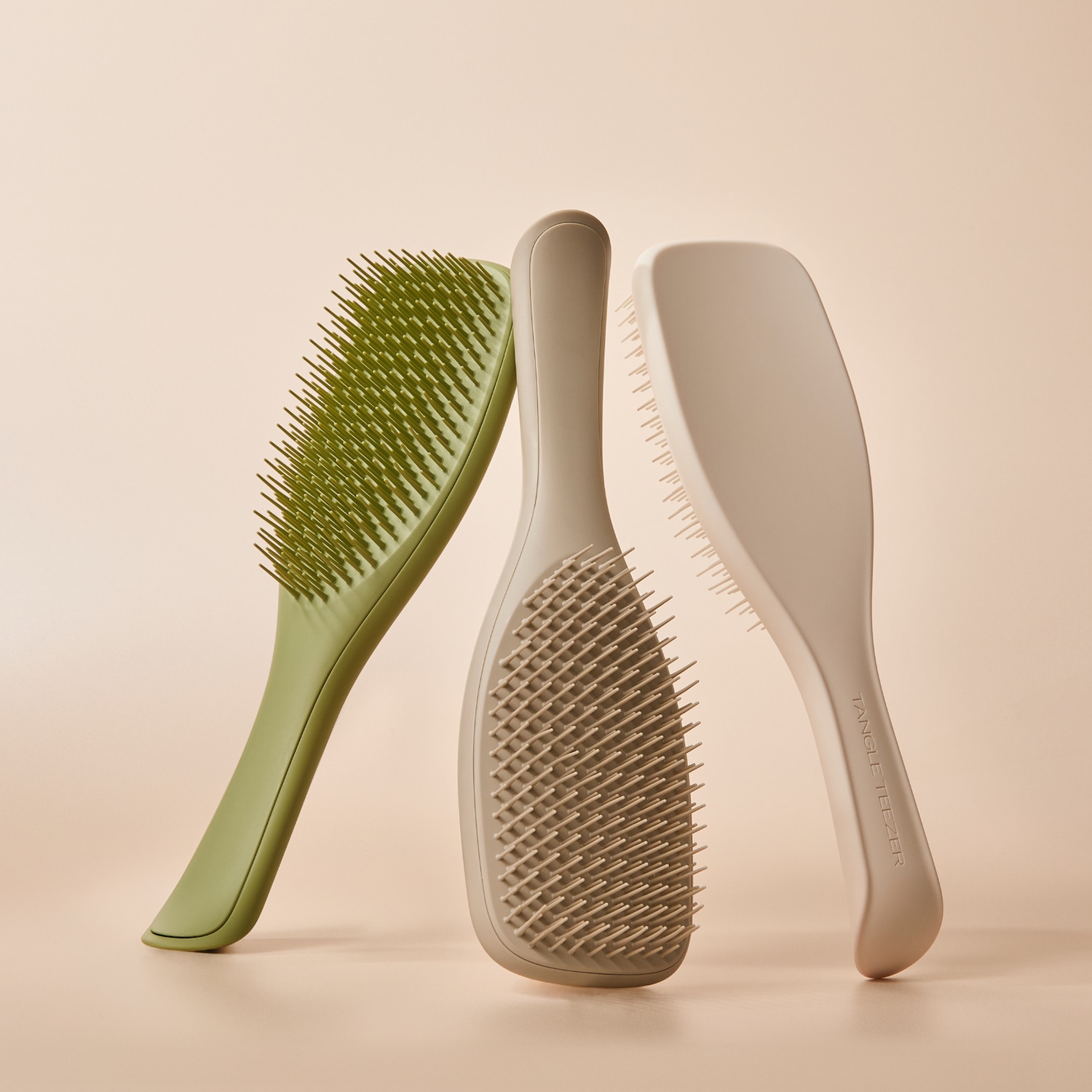 Drei Tangle Teezer Haarbürsten in verschiedenen Grüntönen stehen aufrecht vor einem hellbeigen Hintergrund und zeigen ihre flexiblen Borsten.