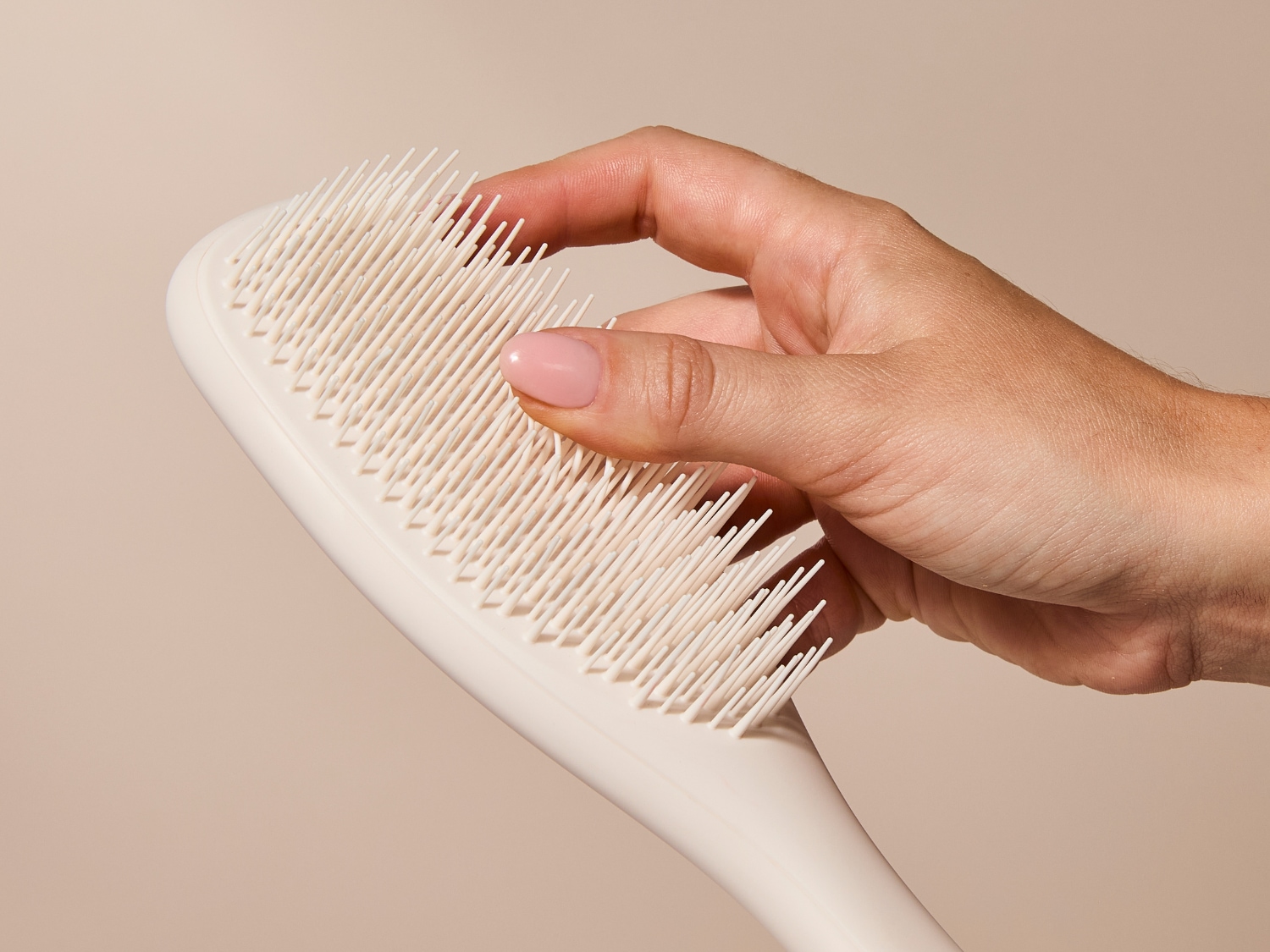 Eine Hand mit rosafarbenen Nägeln hält eine cremefarbene Haarbürste mit weichen Borsten, die sanft über die Kopfhaut gleiten.