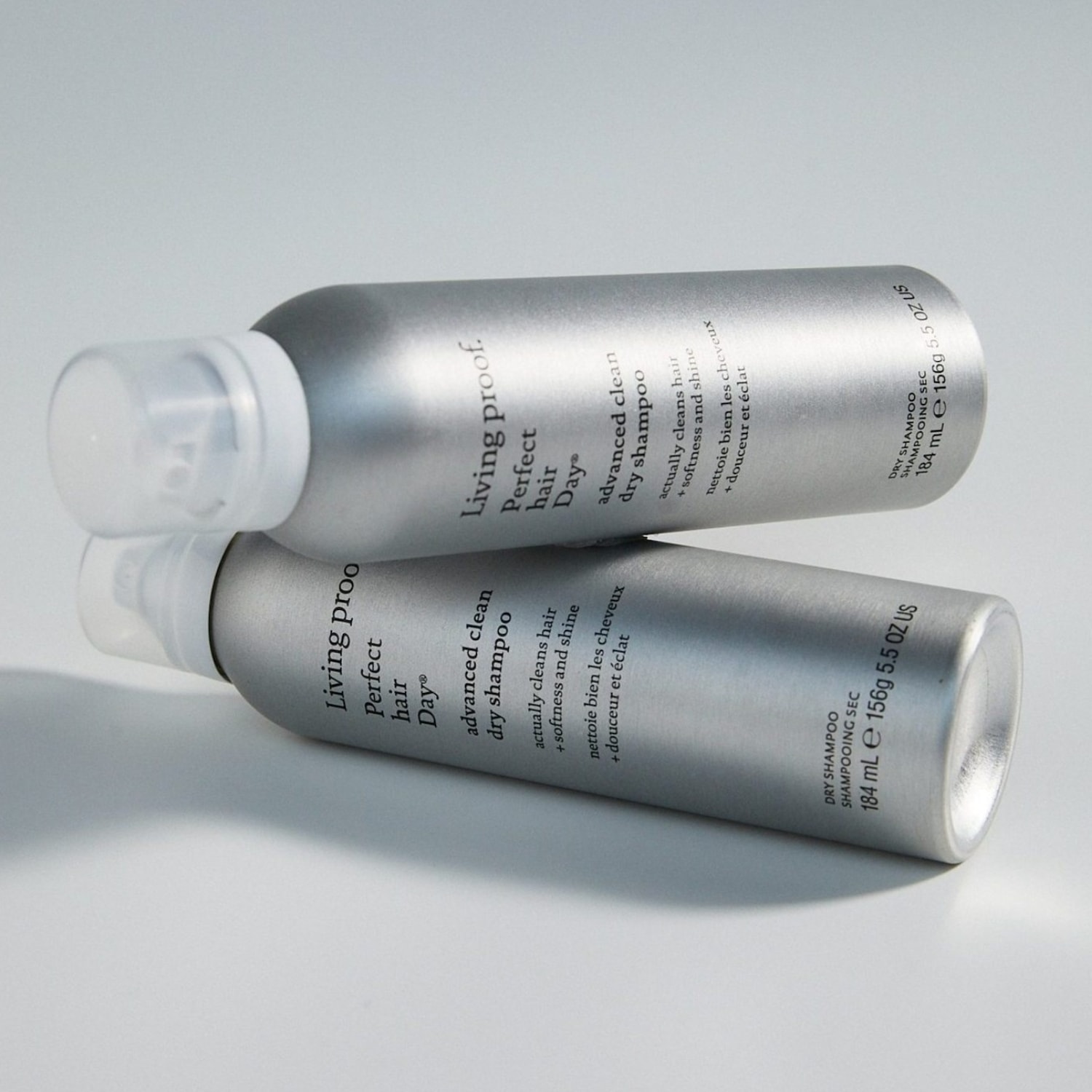 Zwei silberne Flaschen Living Proof Perfect Hair Day Advanced Clean Trockenshampoo liegen auf einem hellgrauen Hintergrund, wobei die obere Flasche leicht über der unteren liegt und beide die Aufschrift „actually cleans hair + softness and shine“ tragen.