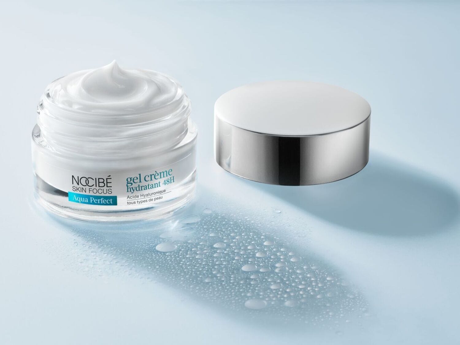 Un pot ouvert de crème hydratante Nocibé Skin Focus Aqua Perfect gel crème hydratant 48H à l'acide hyaluronique pour tous types de peau, avec son couvercle argenté posé à côté, sur un fond bleu clair parsemé de gouttelettes d'eau.