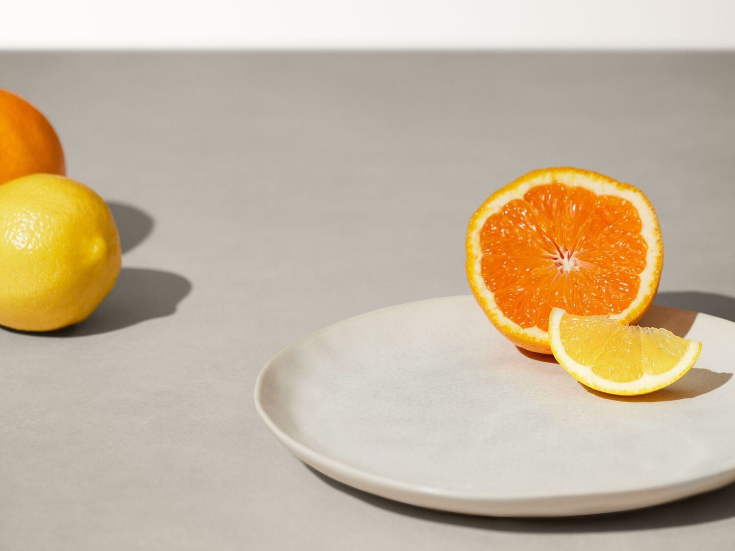 Une orange coupée en deux et un quartier de citron sont posés sur une assiette blanche, avec un citron entier et une orange en arrière-plan, le tout sur une surface grise.