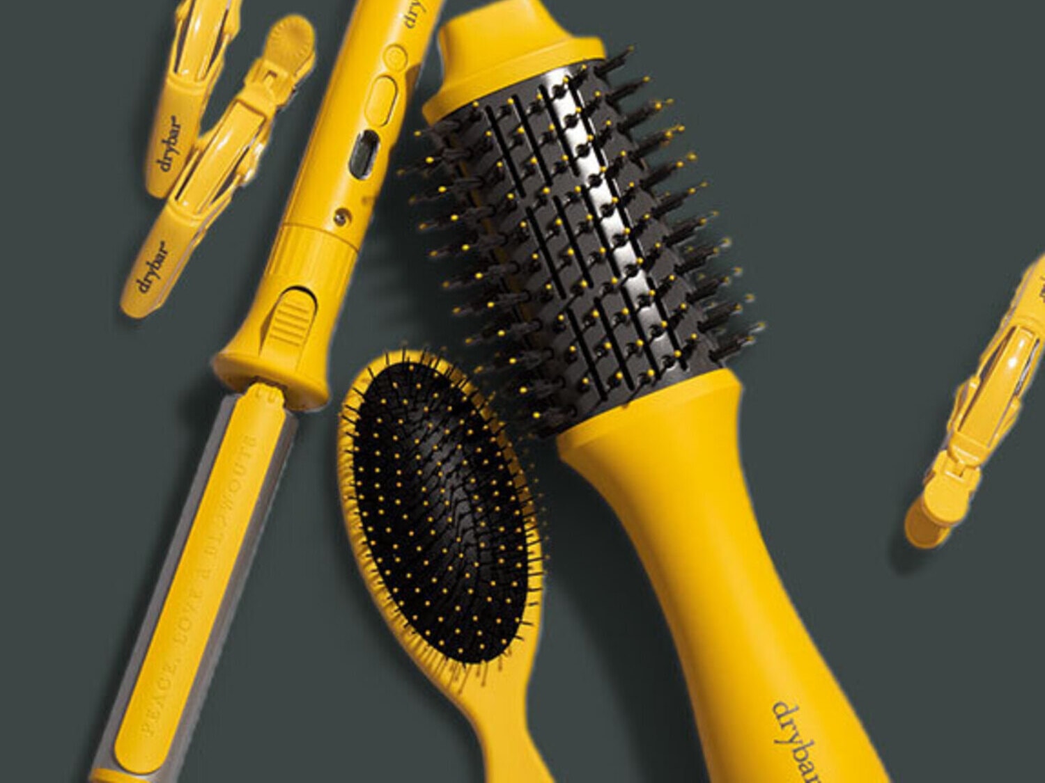 Šiame vaizde matomas „Drybar“ plaukų priežiūros priemonių rinkinys, kuriame yra geltonas plaukų tiesintuvas, du plaukų segtukai, ovalus plaukų šepetys ir apvalus plaukų šepetys, visi su juodais šereliais ir geltonomis rankenomis, išdėstyti ant tamsiai pilko fono.