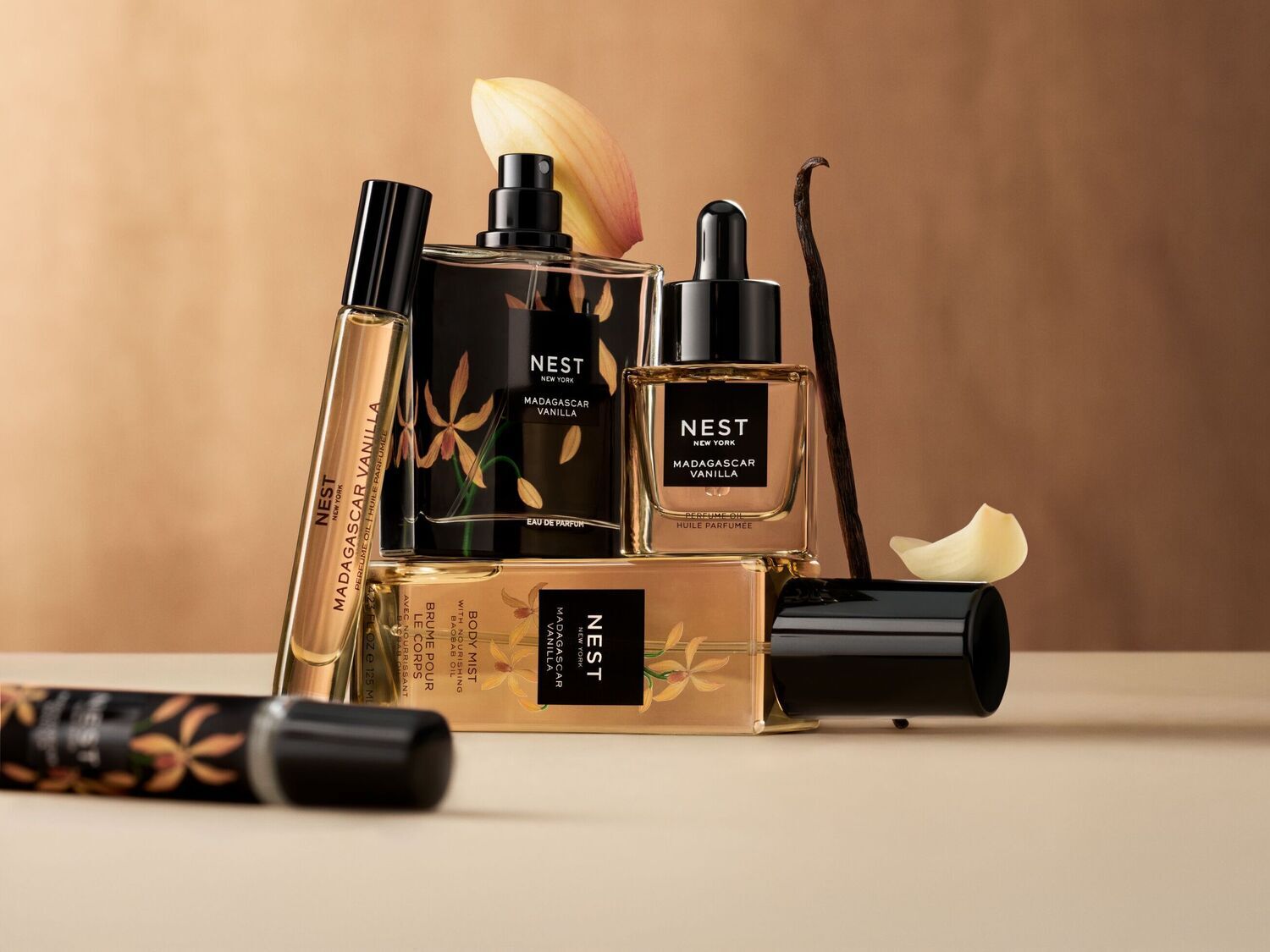 Šiame atvaizde matomas „Nest New York Madagascar Vanilla“ kvapų rinkinys, kuriame yra kvepalų aliejus, kūno dulksna ir parfumuotas vanduo, išdėstyti ant smėlio spalvos paviršiaus su vanilės ankštimis ir žiedlapiais, sukuriant prabangų ir viliojantį vaizdą.