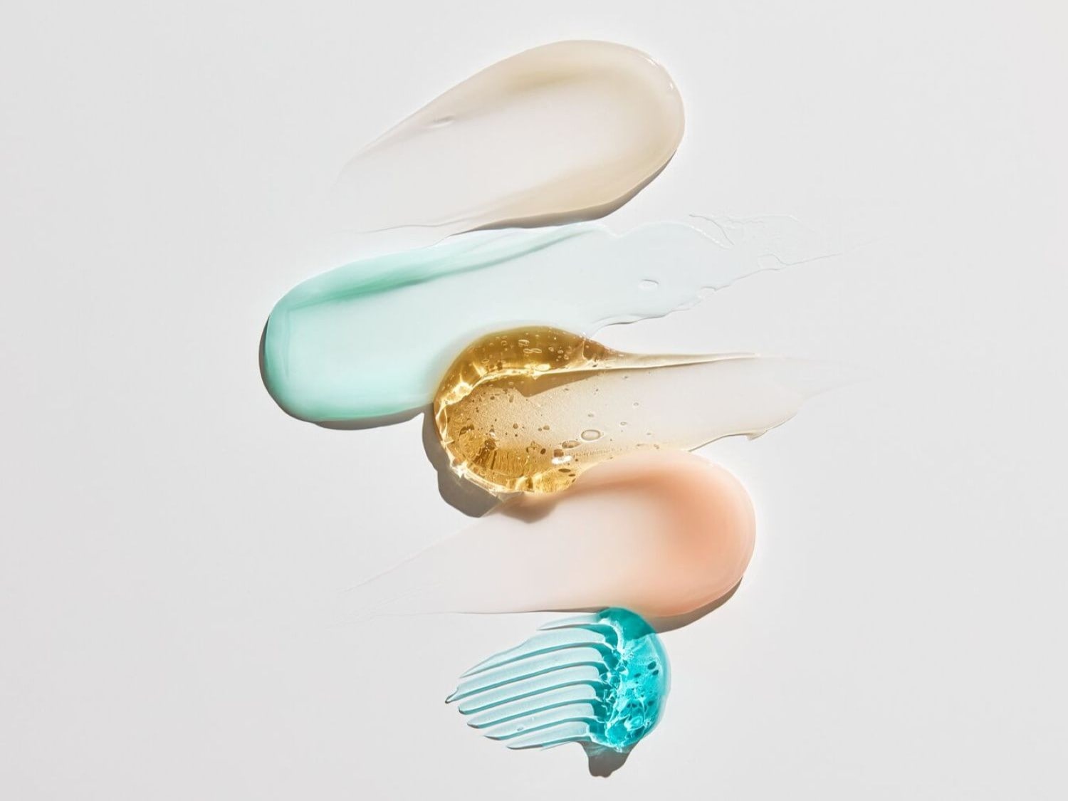 Una serie di strisce di prodotti di bellezza di diversi colori e texture, tra cui un gel trasparente, una crema bianca, una lozione verde acqua, un siero dorato e un gel blu, sono disposte su uno sfondo bianco.