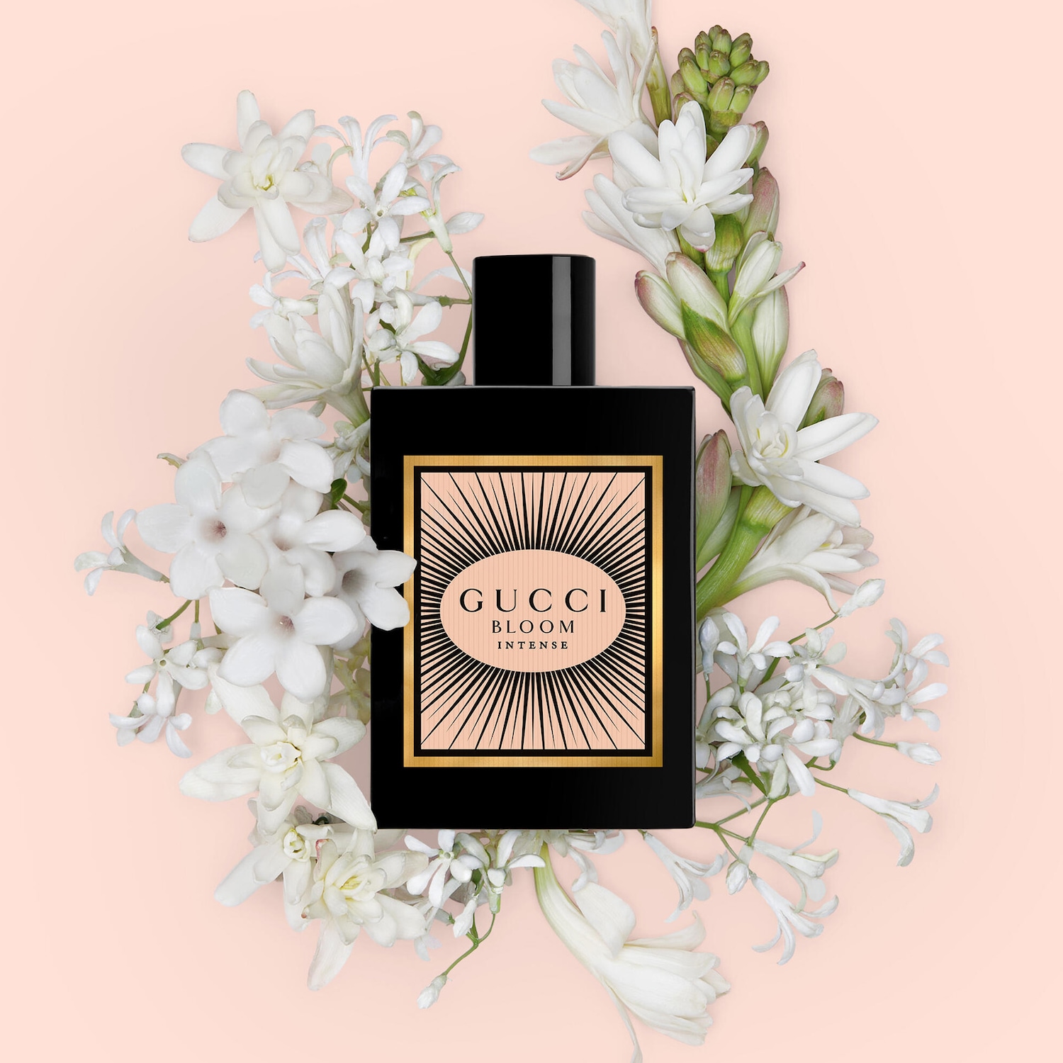 Černý flakon parfému Gucci Bloom Intense s broskvovým štítkem se zlatým orámováním a nápisem Gucci Bloom Intense, obklopený bílými květy na světle růžovém pozadí.