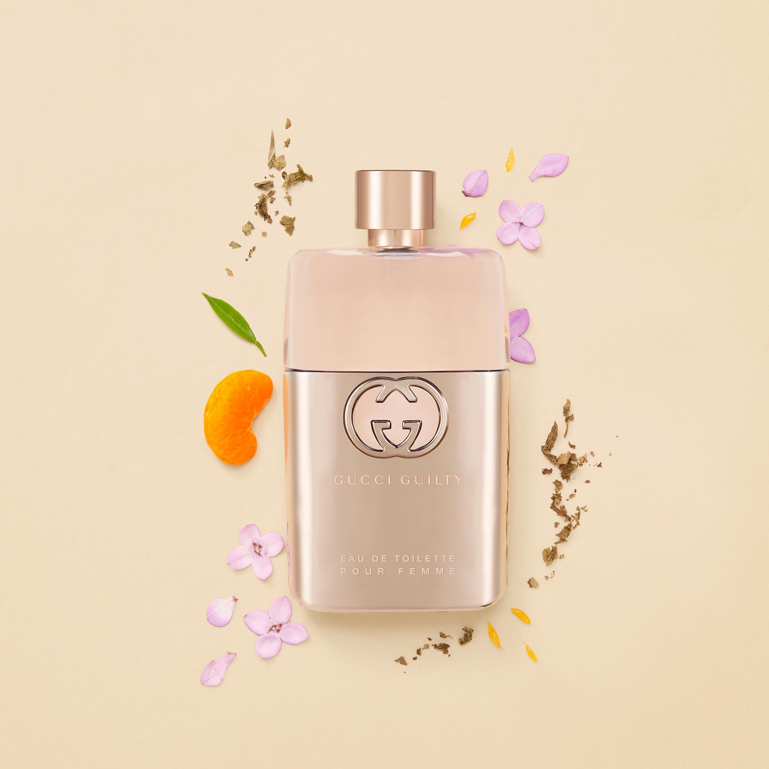 Parfém Gucci Guilty Eau de Toilette Pour Femme ve zlatém flakonu s logem Gucci uprostřed, obklopený okvětními lístky a bylinkami, leží na béžovém pozadí.