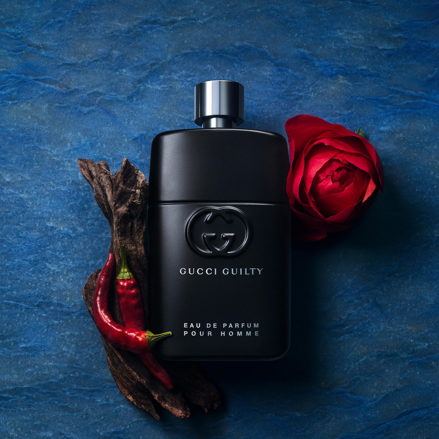 Gucci Guilty Pour Homme Eau de Parfum je prezentován v elegantním černém flakonu s kovovým uzávěrem, doplněný červenou růží, chilli papričkami a kousky dřeva na modrém pozadí.