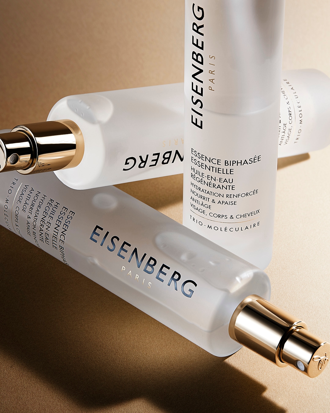 Tres botellas de Eisenberg Paris Essence Biphasee Essentielle, un aceite en agua regenerador para rostro, cuerpo y cabello, se muestran sobre una superficie dorada, destacando su diseño elegante y su promesa de hidratación y nutrición.