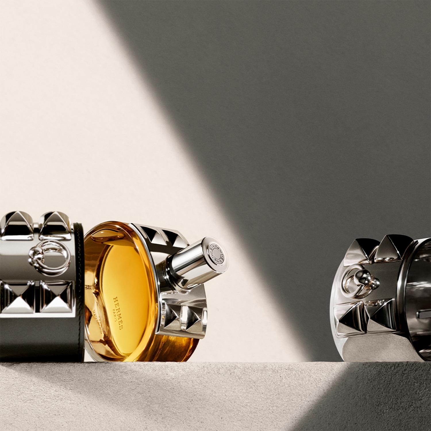Le parfum Hermès est présenté dans un flacon rond en verre transparent avec un liquide ambré, orné d'un bracelet en cuir noir clouté d'argent, posé sur une surface texturée.