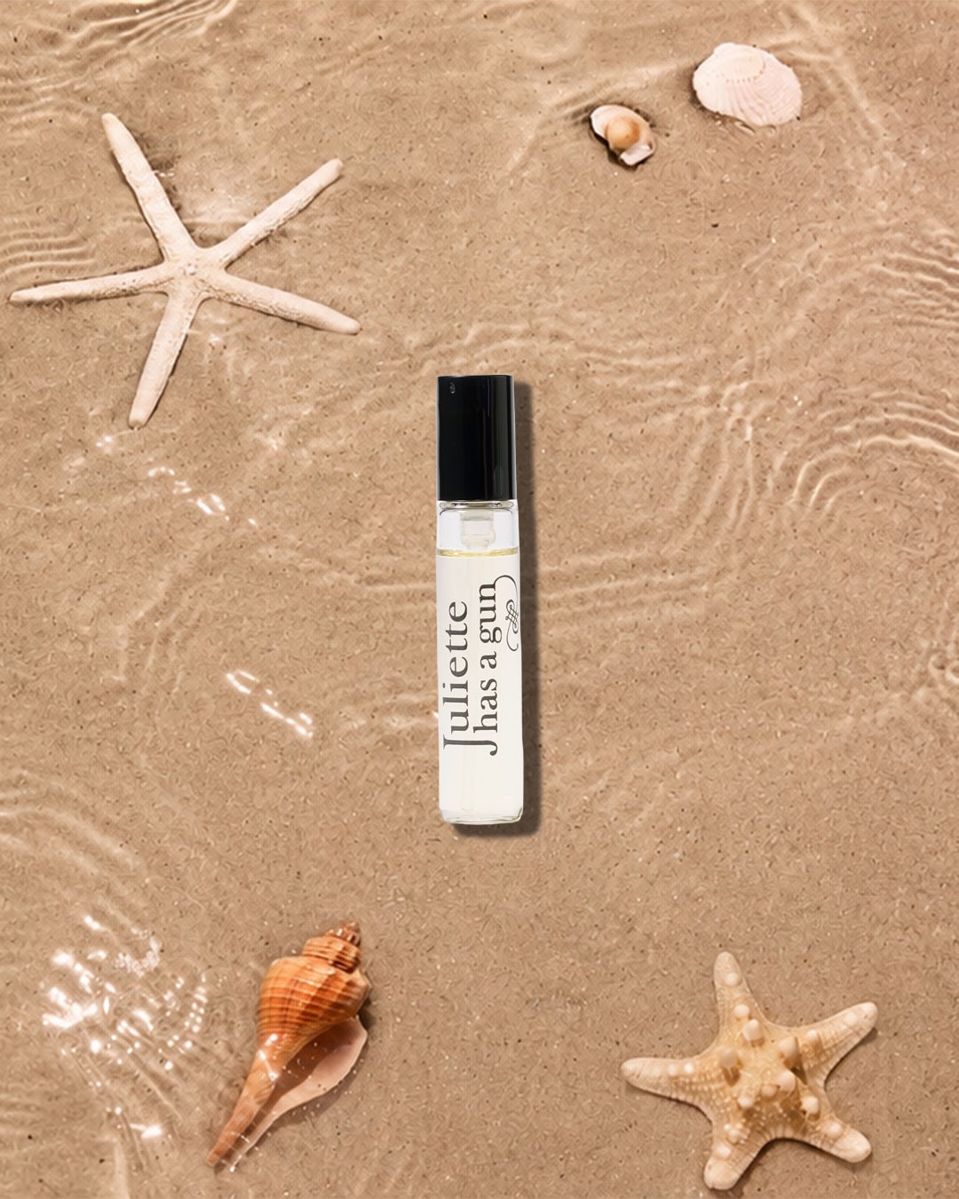 Ein Juliette Has a Gun Parfum-Rollerball liegt auf nassem Sand, umgeben von Seesternen und Muscheln, mit leichtem Wellengang, der eine sommerliche Strandatmosphäre schafft.