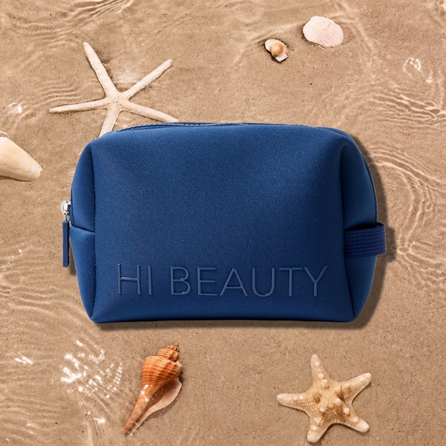 Eine blaue Kosmetiktasche mit der Aufschrift „HI BEAUTY“ liegt auf sandigem Untergrund, umgeben von Muscheln und Seesternen, mit leichtem Wellengang im Hintergrund.