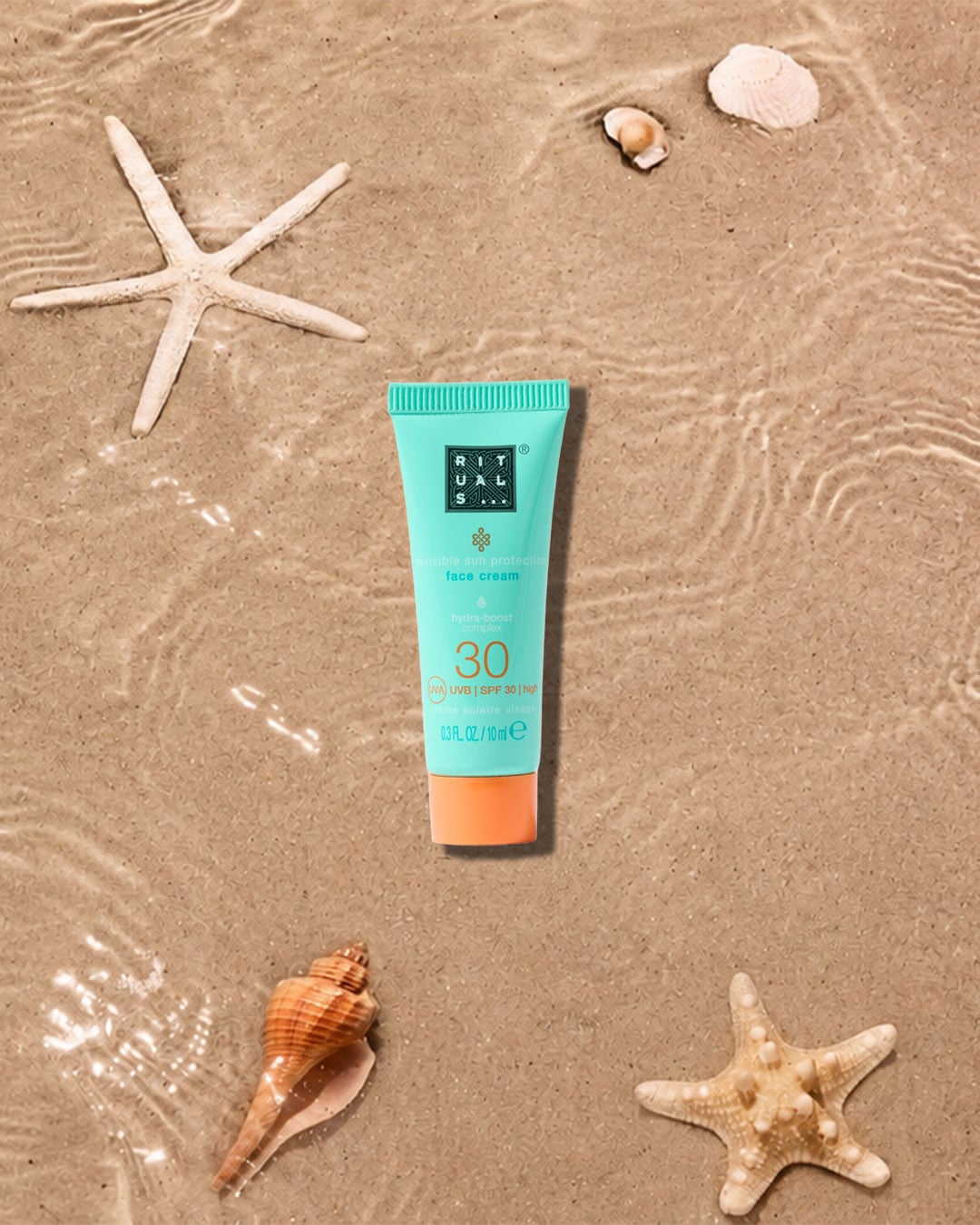 Die RITUALS Invisible Sun Protection Face Cream SPF 30 in einer türkisfarbenen Tube mit orangefarbener Kappe liegt auf nassem Sand, umgeben von Muscheln und Seesternen.