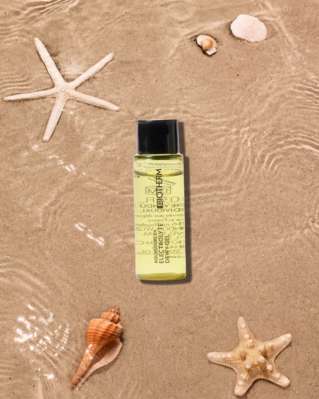 Das Biotherm Aquasource Electrolyte Dewy Gel in einer transparenten Flasche mit schwarzem Deckel, umgeben von Seesternen und Muscheln auf sandigem Untergrund mit Wasserspiegelungen, bietet intensive Feuchtigkeit für die Haut.