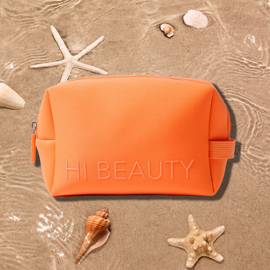 Eine leuchtend orangefarbene Kosmetiktasche mit der Aufschrift "HI BEAUTY" liegt auf nassem Sand, umgeben von Muscheln und Seesternen.