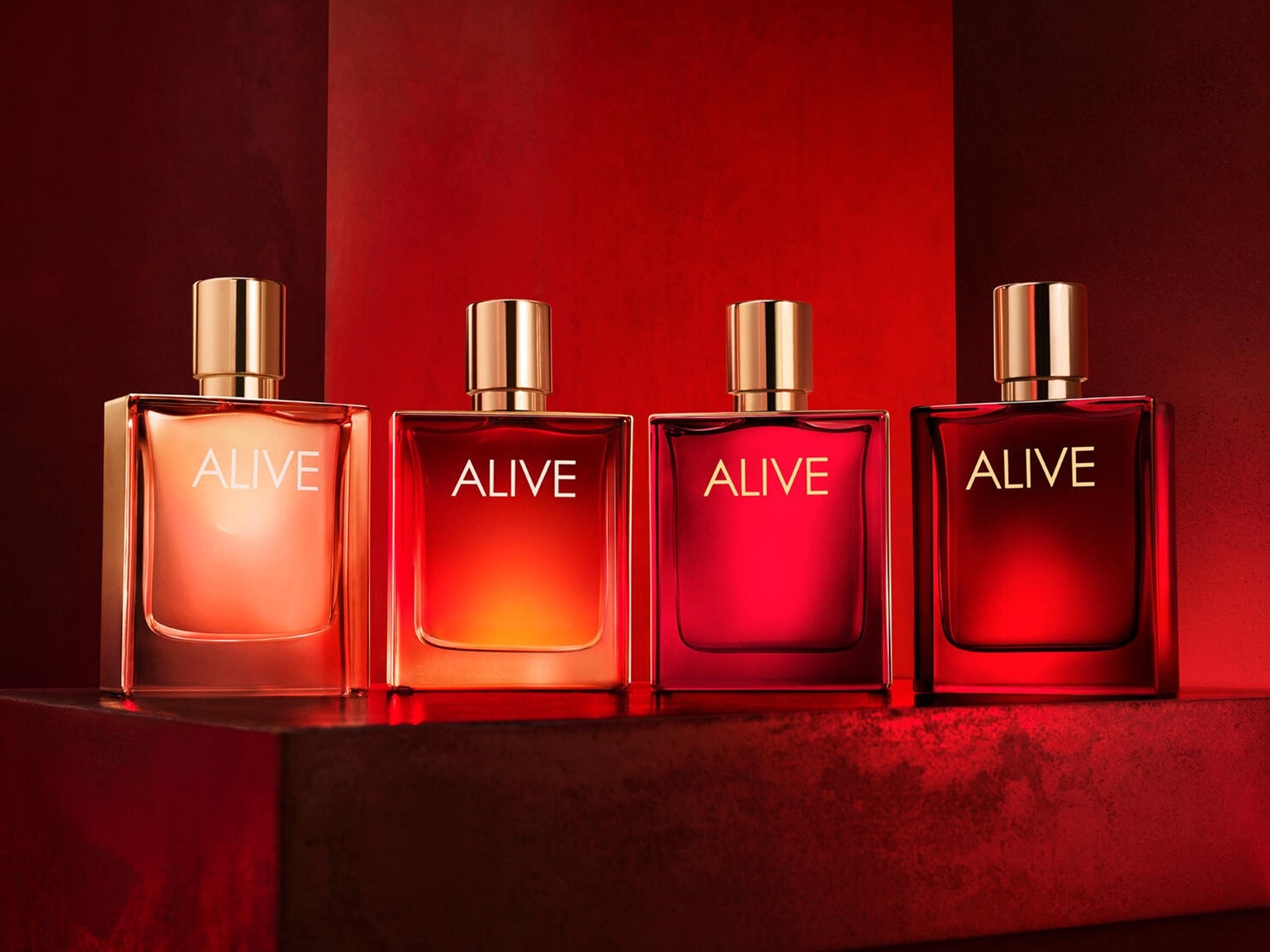 Patru sticle de parfum "Alive" cu capace aurii, aranjate pe o suprafață roșie, cu nuanțe de la portocaliu deschis la roșu închis, creând un efect de gradient.