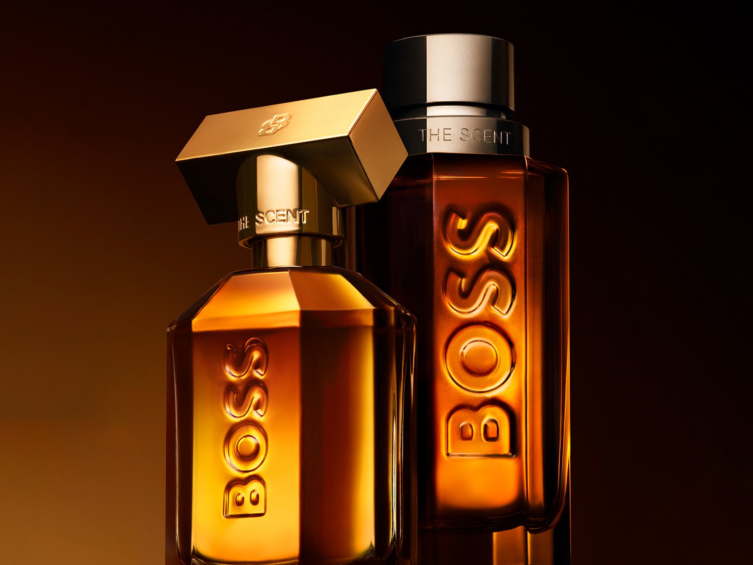 Două sticle de parfum Hugo Boss The Scent, una mai mică în față și una mai mare în spate, ambele cu lichid auriu-chihlimbar și capace aurii, pe un fundal maro închis.