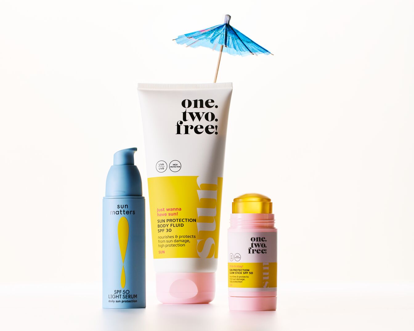 Trijų saulės apsaugos priemonių rinkinys: mėlynas „Sun Matters SPF 50 Light Serum“, baltas „One.Two.Free! Sun Protection Body Fluid SPF 30“ ir rožinis „One.Two.Free! Sun Protection Glow Stick SPF 50“, papuoštas mažu mėlynu skėčiu.