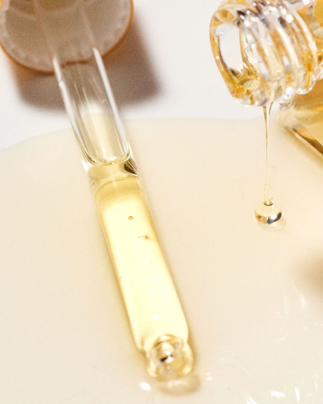 Un sérum doré est versé d'une pipette en verre sur une surface claire, avec une goutte suspendue au flacon, illustrant la texture riche et l'application précise du produit.