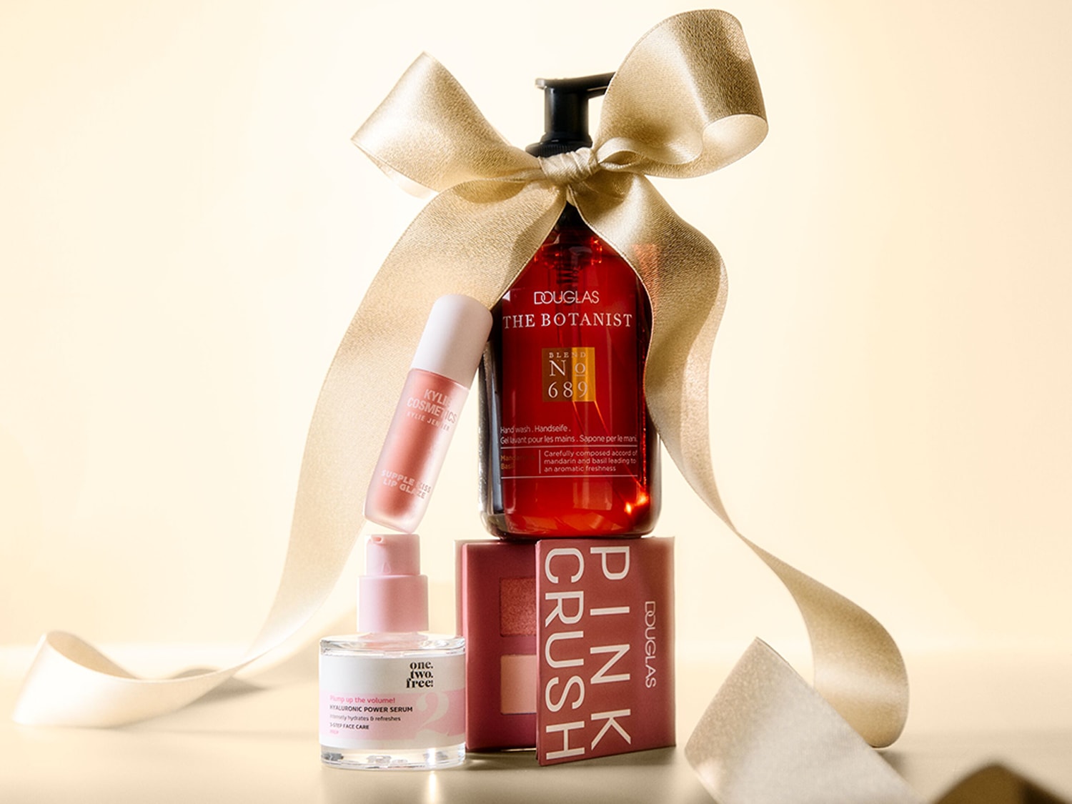 Ein festliches Geschenkset mit einem Douglas The Botanist Handwaschmittel in einer roten Flasche, einem Kylie Cosmetics Lip Glaze, einem One.Two.Free. Hyaluronic Power Serum und einer Douglas Pink Crush Lidschattenpalette, alles mit einem goldenen Band verziert.