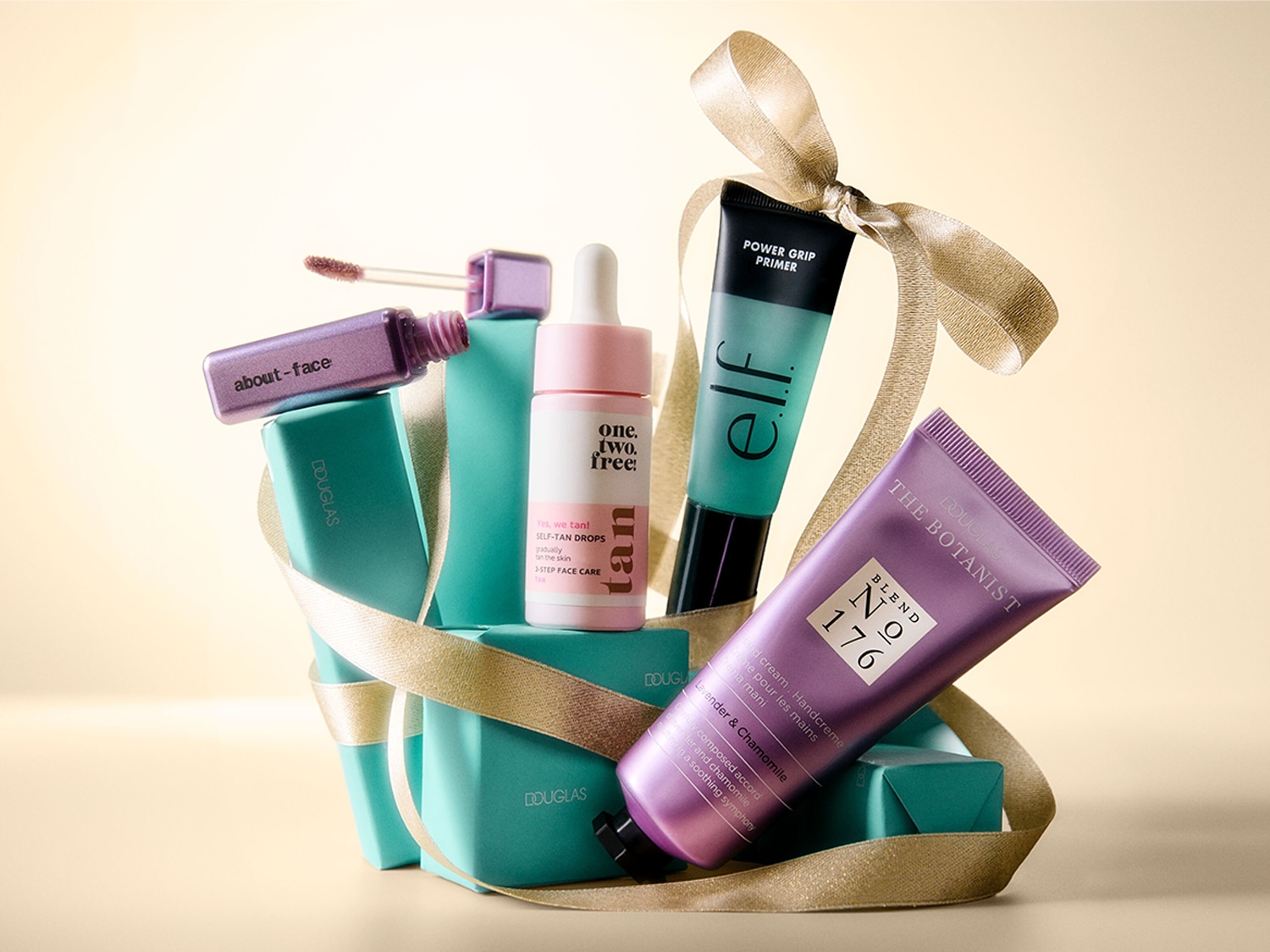 Ein festliches Arrangement aus Beauty-Produkten, darunter ein lila Lipgloss von "about-face", ein Selbstbräuner von "one.two.free!", ein grüner "elf" Power Grip Primer und eine lila Handcreme von "DOUGLAS THE BOTANIST", allesamt mit einem goldenen Band umwickelt und auf türkisfarbenen Geschenkboxen platziert.