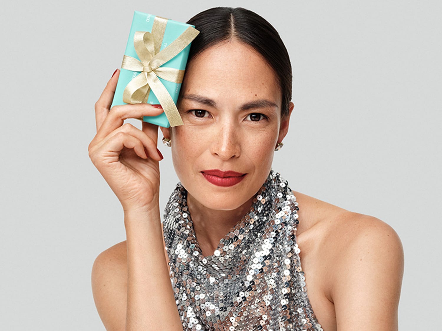 Eine Frau mit einem glitzernden silbernen Oberteil hält ein kleines, türkisfarbenes Geschenk mit einer goldenen Schleife neben ihrem Gesicht.