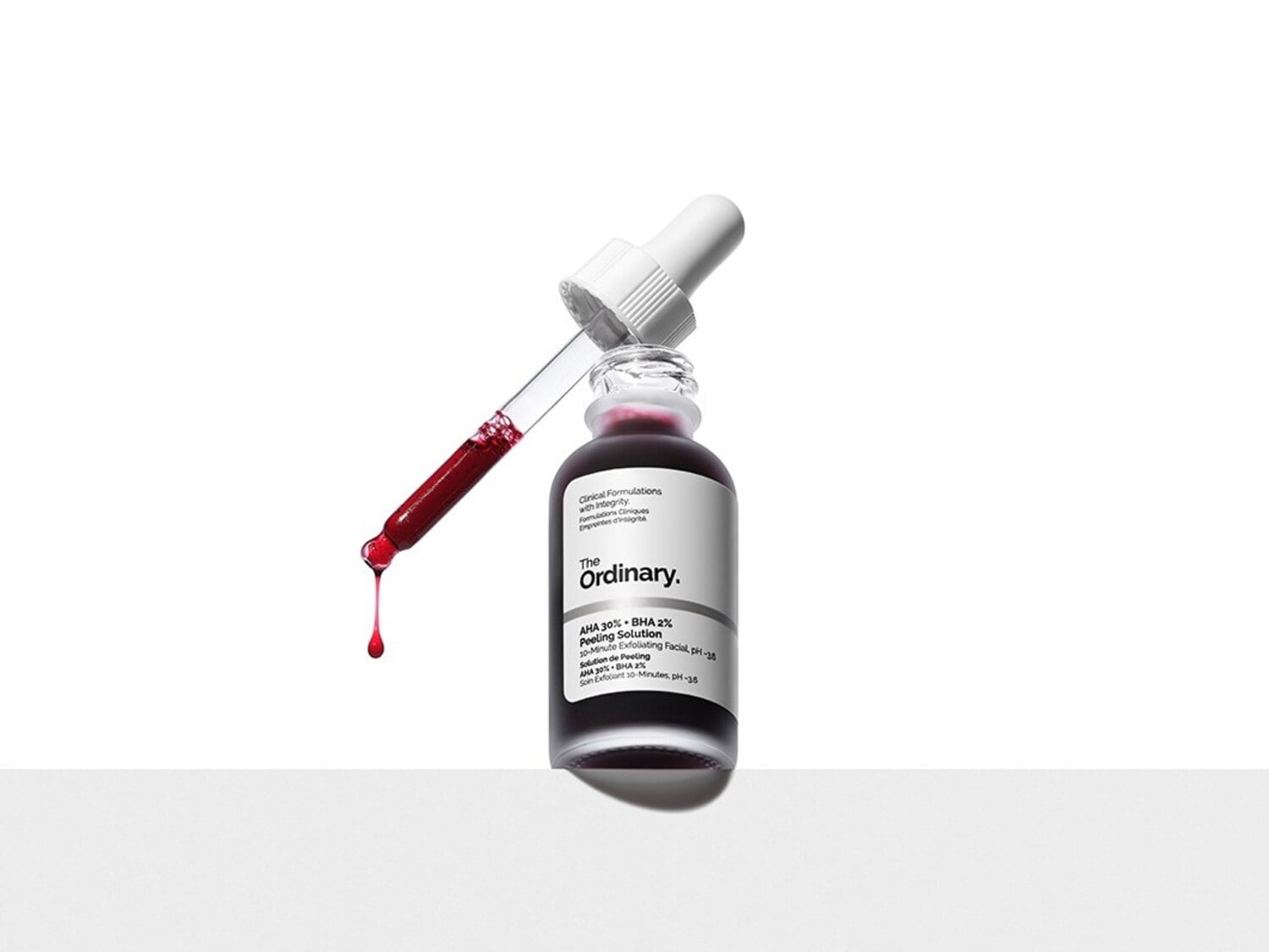 Le sérum The Ordinary AHA 30% + BHA 2% Peeling Solution est présenté dans une bouteille en verre transparent avec une étiquette blanche, un compte-gouttes blanc et un liquide rouge foncé, avec une goutte de sérum tombant du compte-gouttes.