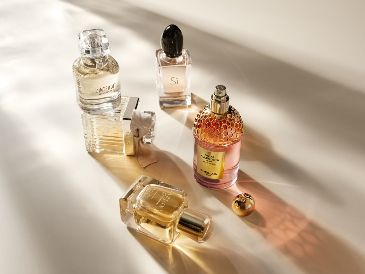 Pięć eleganckich butelek perfum, w tym L'Interdit Givenchy, Si, Chloe, Aqua Allegoria Forte Rosa Rossa Guerlain i Jardin, Empreinte Amour, jest pięknie oświetlonych, tworząc grę światła i cienia.