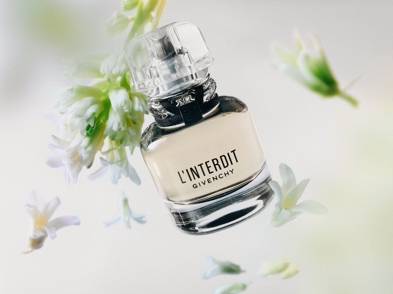 Flakon perfum L'Interdit Givenchy unosi się w powietrzu, otoczony delikatnymi białymi kwiatami, co podkreśla jego świeży i elegancki charakter.