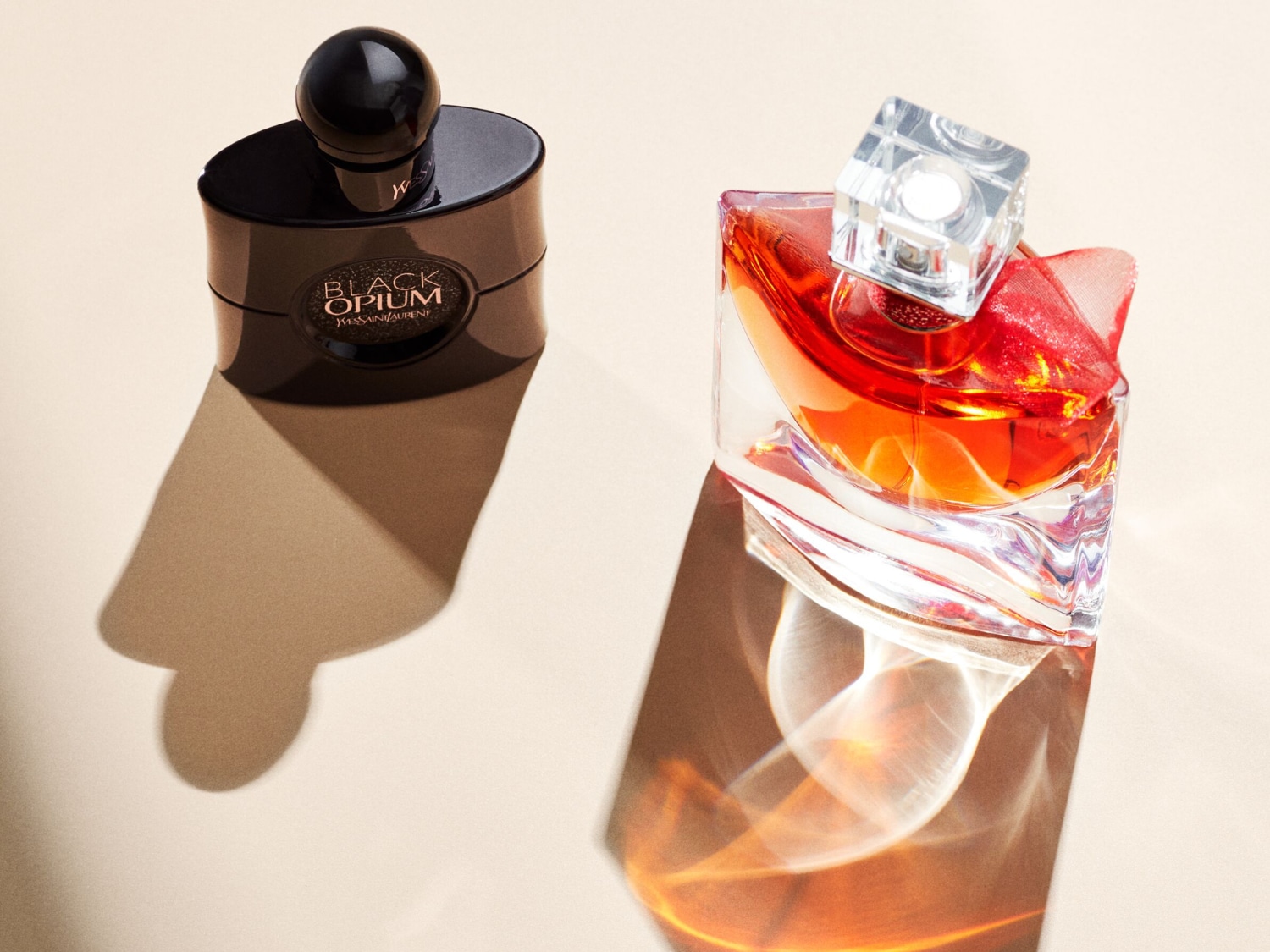 Dwa flakony perfum, jeden czarny z napisem "Black Opium Yves Saint Laurent", a drugi przezroczysty z pomarańczowym płynem i czerwoną kokardką, stoją na beżowym tle, rzucając długie cienie.