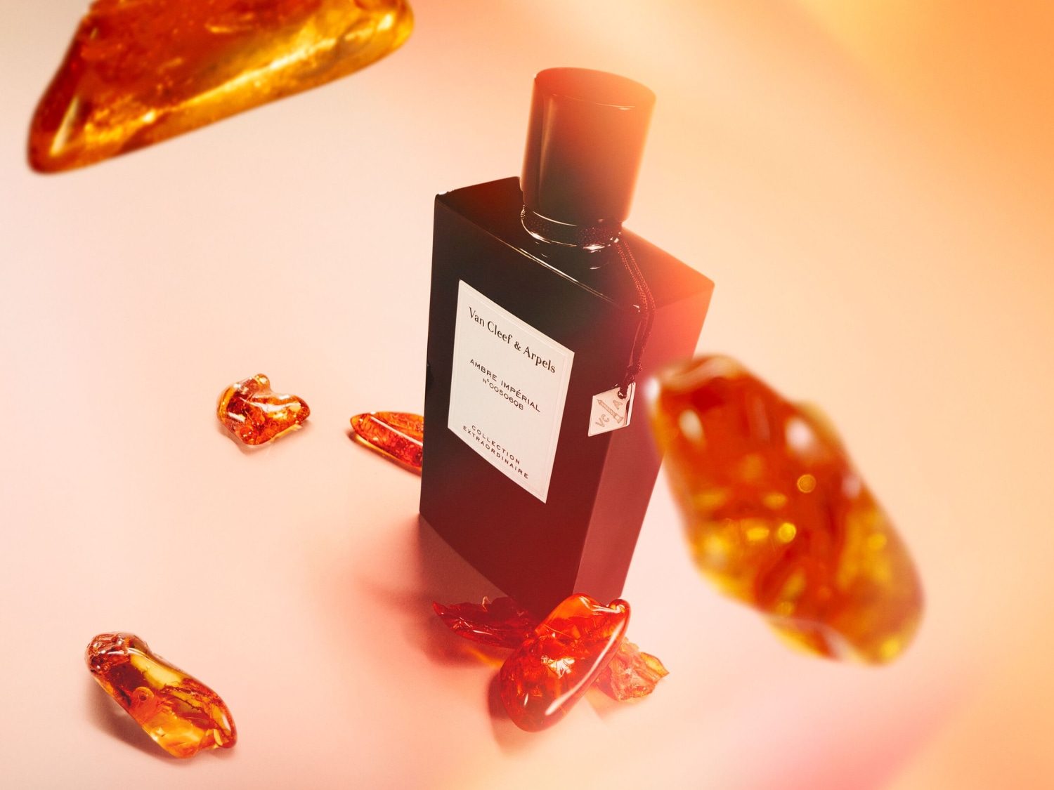 Flakon perfum Van Cleef & Arpels Ambre Impérial z kolekcji Collection Extraordinaire, otoczony bursztynowymi kamieniami, prezentuje się na tle w odcieniach brzoskwini i pomarańczy.