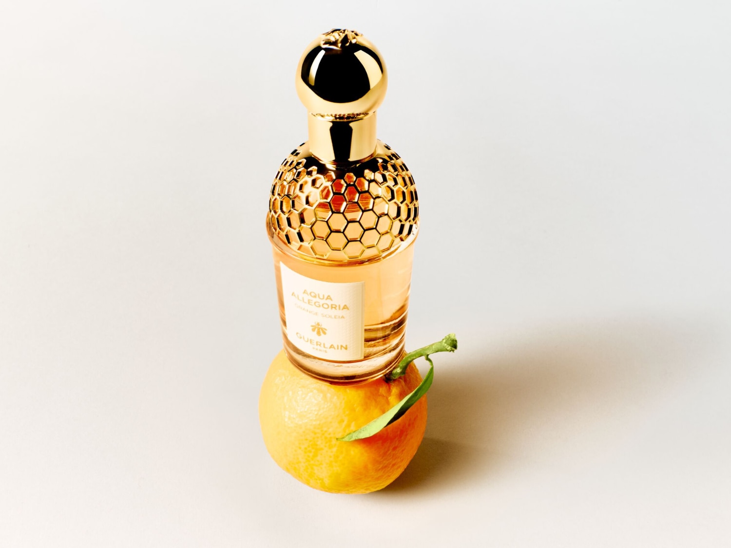 Perfumy Aqua Allegoria Orange Soleia od Guerlain w eleganckiej butelce ze złotym korkiem i miodowym wzorem, stojące na świeżej pomarańczy z zielonym listkiem, na białym tle.