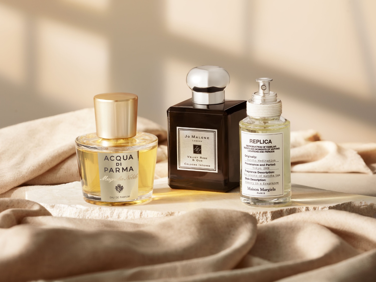 Trzy butelki perfum, w tym Acqua di Parma Magnolia Nobile z żółtym płynem i złotą nakrętką, Jo Malone Velvet Rose & Oud w ciemnej butelce ze srebrną nakrętką oraz Maison Margiela Replica Matcha Meditation z jasnym płynem i srebrnym atomizerem, stoją na beżowym tle z pofałdowanym materiałem.