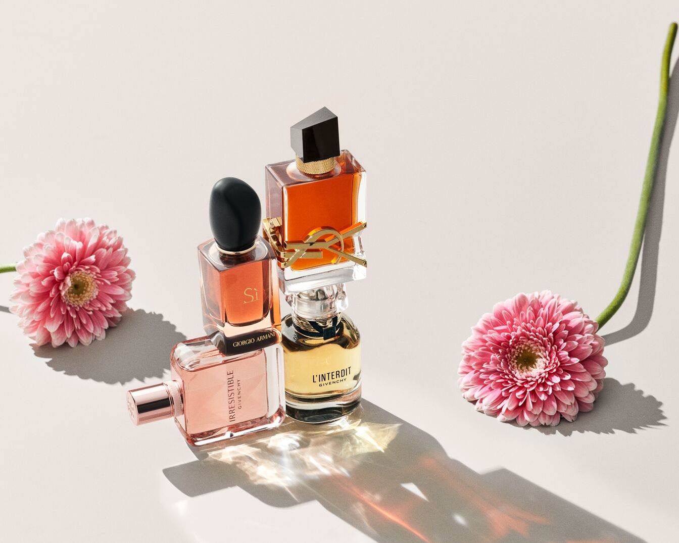 Cztery butelki perfum w różnych odcieniach, od jasnoróżowego po bursztynowy, ułożone są na jasnym tle obok dwóch różowych gerberów, tworząc elegancką kompozycję.