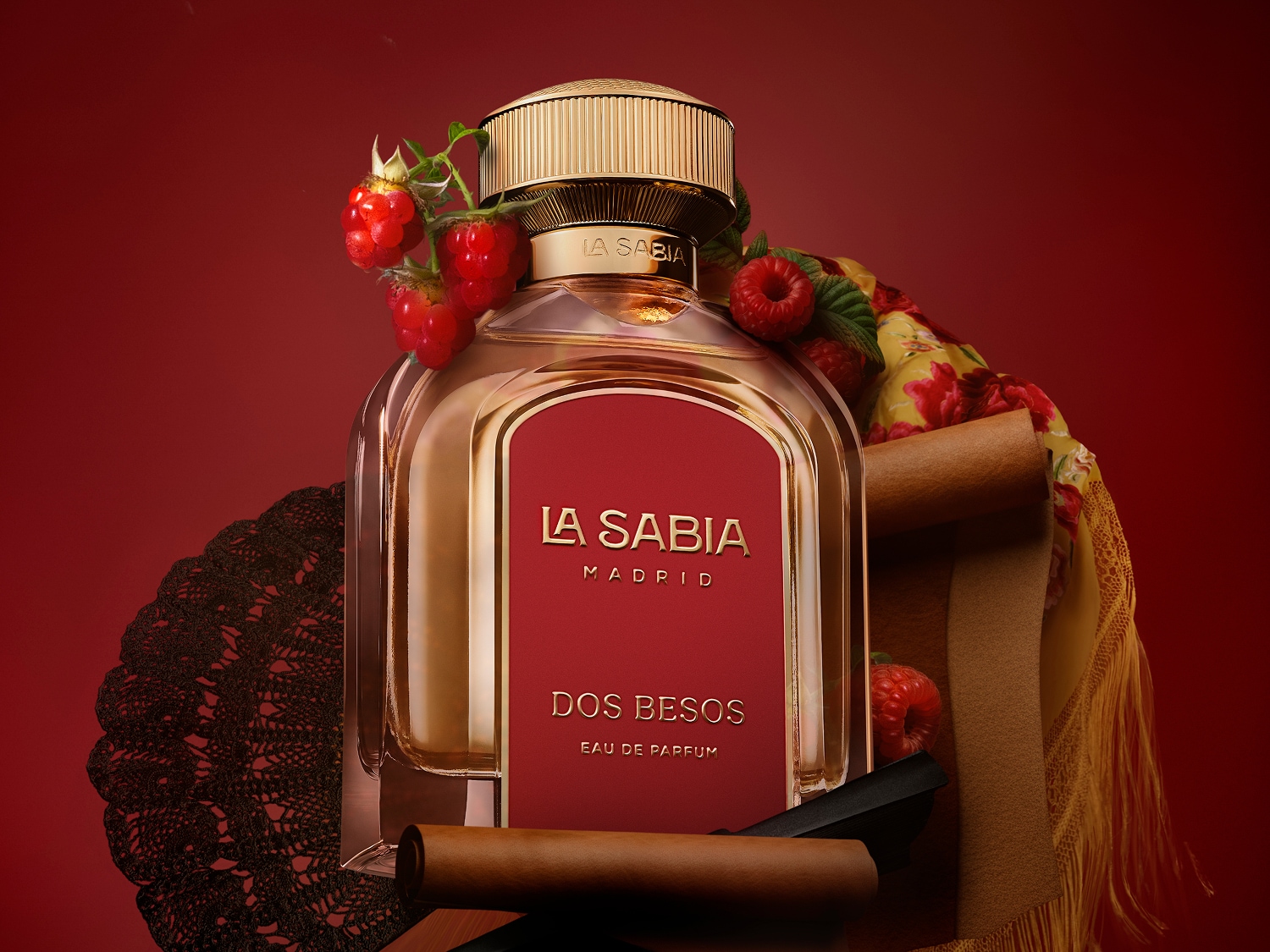 Flakon perfum La Sabia Madrid Dos Besos Eau de Parfum z czerwoną etykietą i złotą nakrętką, ozdobiony malinami, czarną koronką i zwiniętym materiałem, na tle w odcieniu czerwieni.