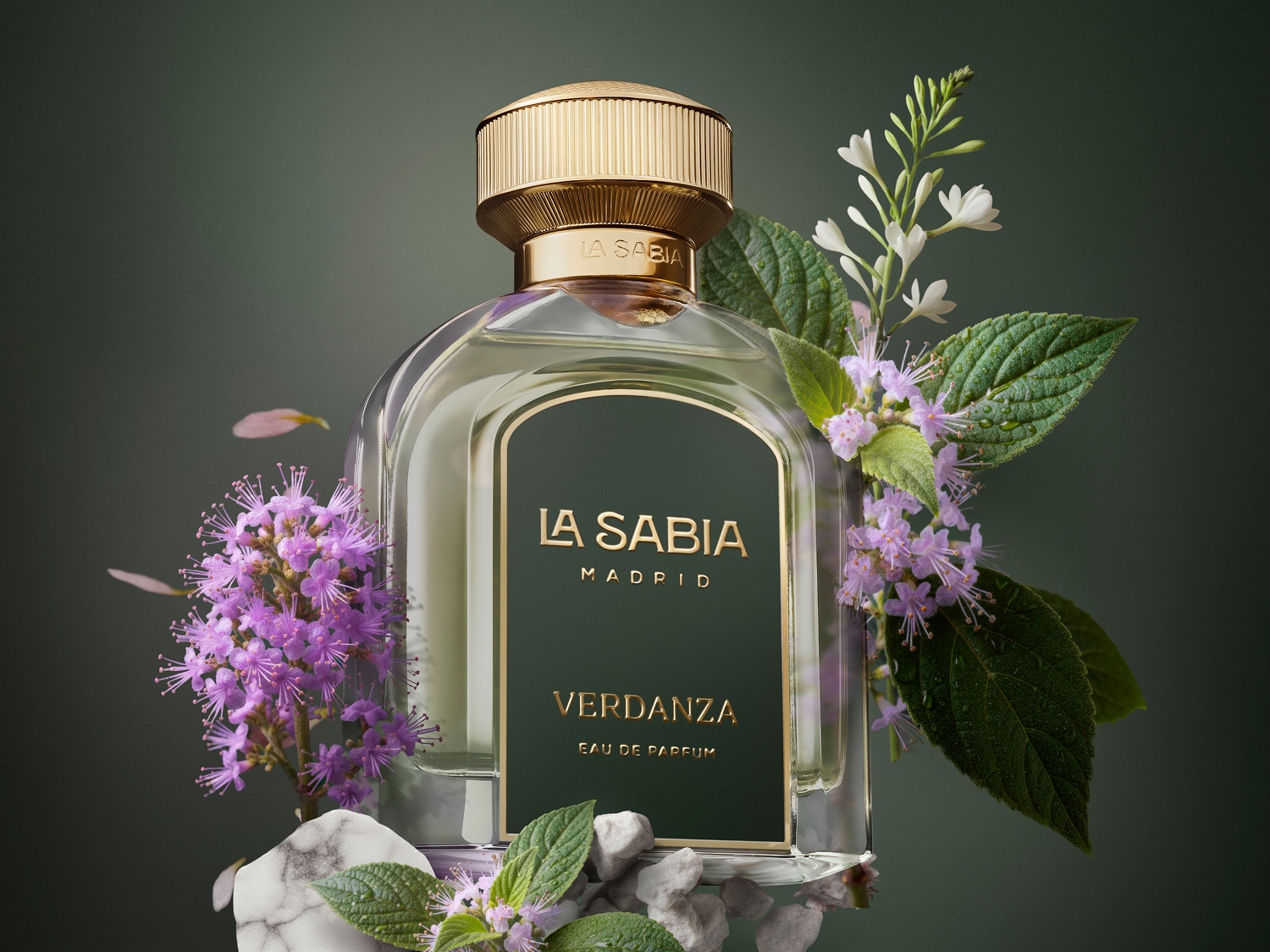 Flakon perfum La Sabia Verdanza Eau de Parfum z zieloną etykietą i złotą nakrętką, otoczony fioletowymi i białymi kwiatami oraz zielonymi liśćmi, spoczywa na marmurowym postumencie z małymi kamykami.
