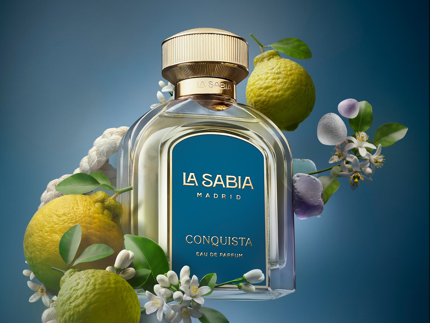 Flakon perfum La Sabia Conquista Eau de Parfum z elegancką złotą nakrętką, otoczony świeżymi cytrusami, białymi kwiatami i ozdobnymi kamieniami, na tle błękitnego nieba.