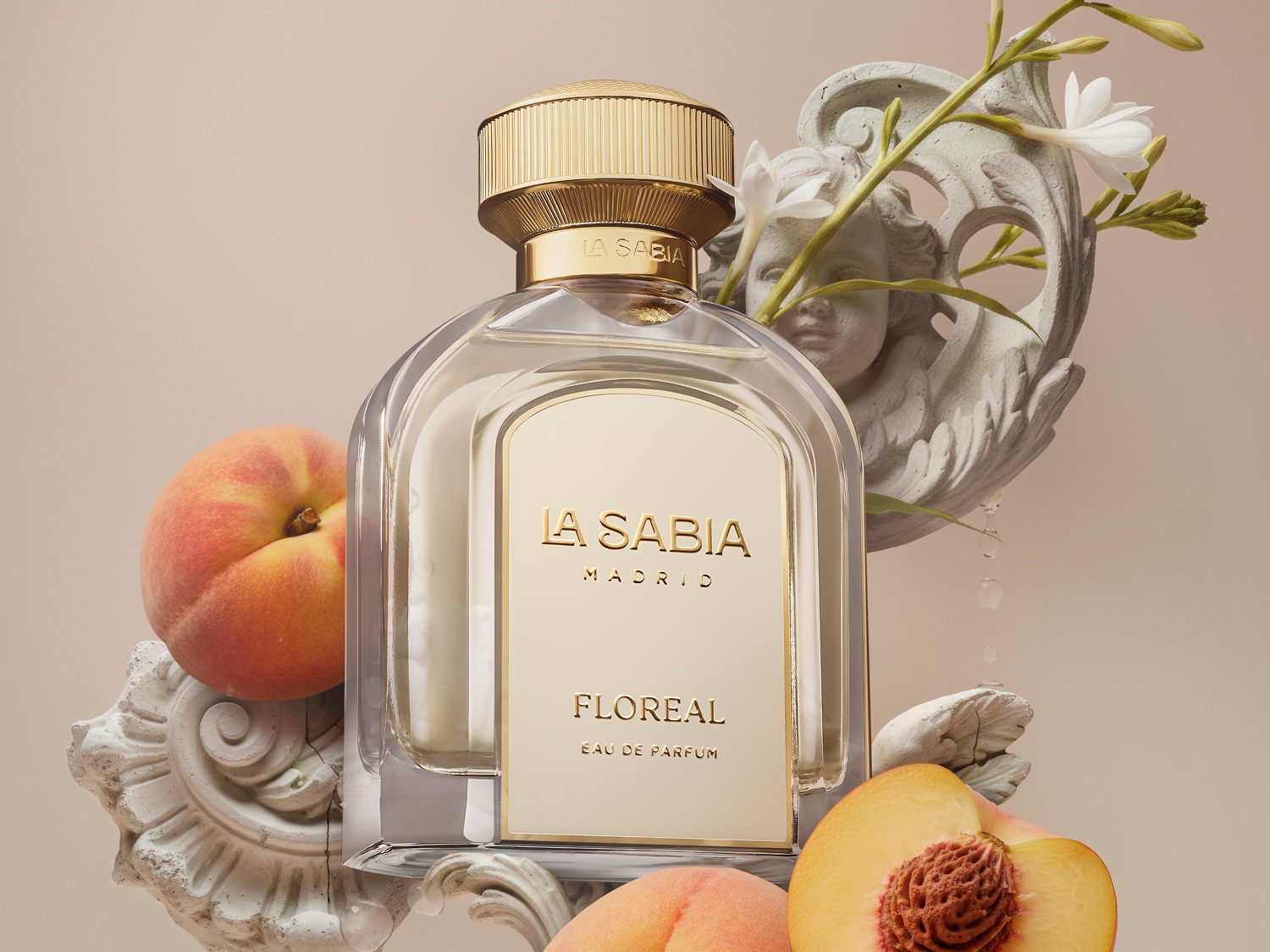 Perfumy La Sabia Floreal Eau de Parfum w przezroczystej butelce ze złotą nakrętką, otoczone brzoskwiniami i rzeźbionymi elementami, z delikatnymi białymi kwiatami.