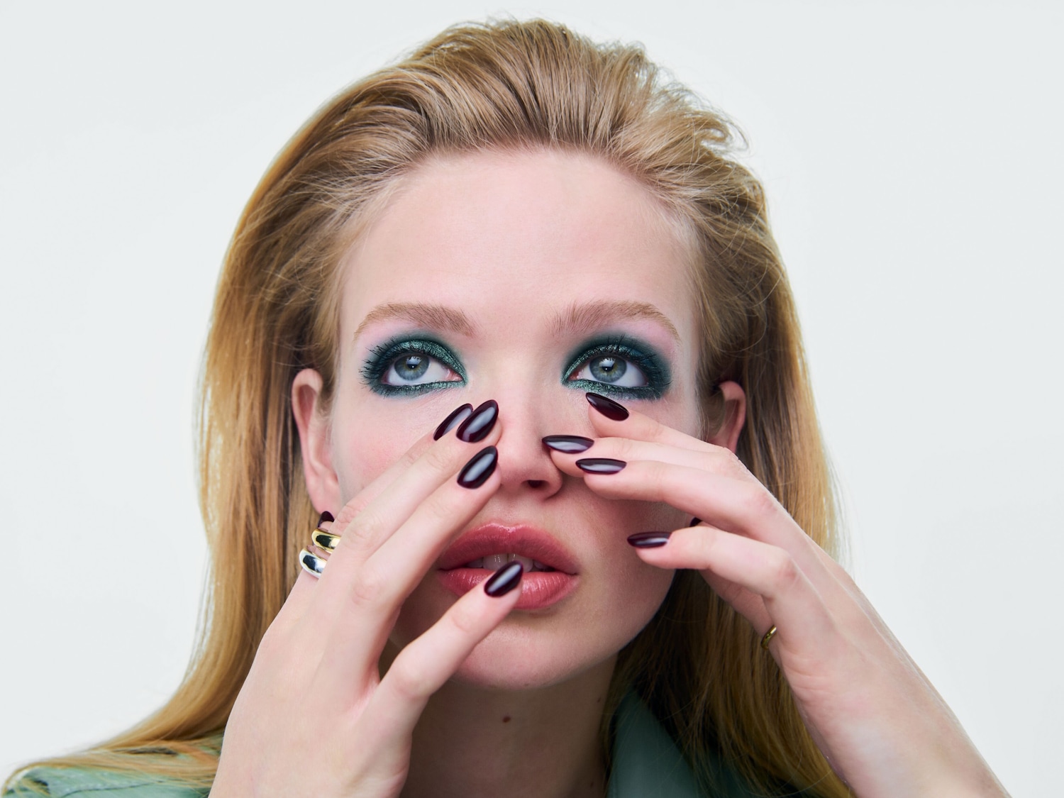 Una donna con lunghi capelli biondi e un trucco occhi verde smeraldo intenso, con unghie laccate di bordeaux lucido, si tocca il viso con entrambe le mani.