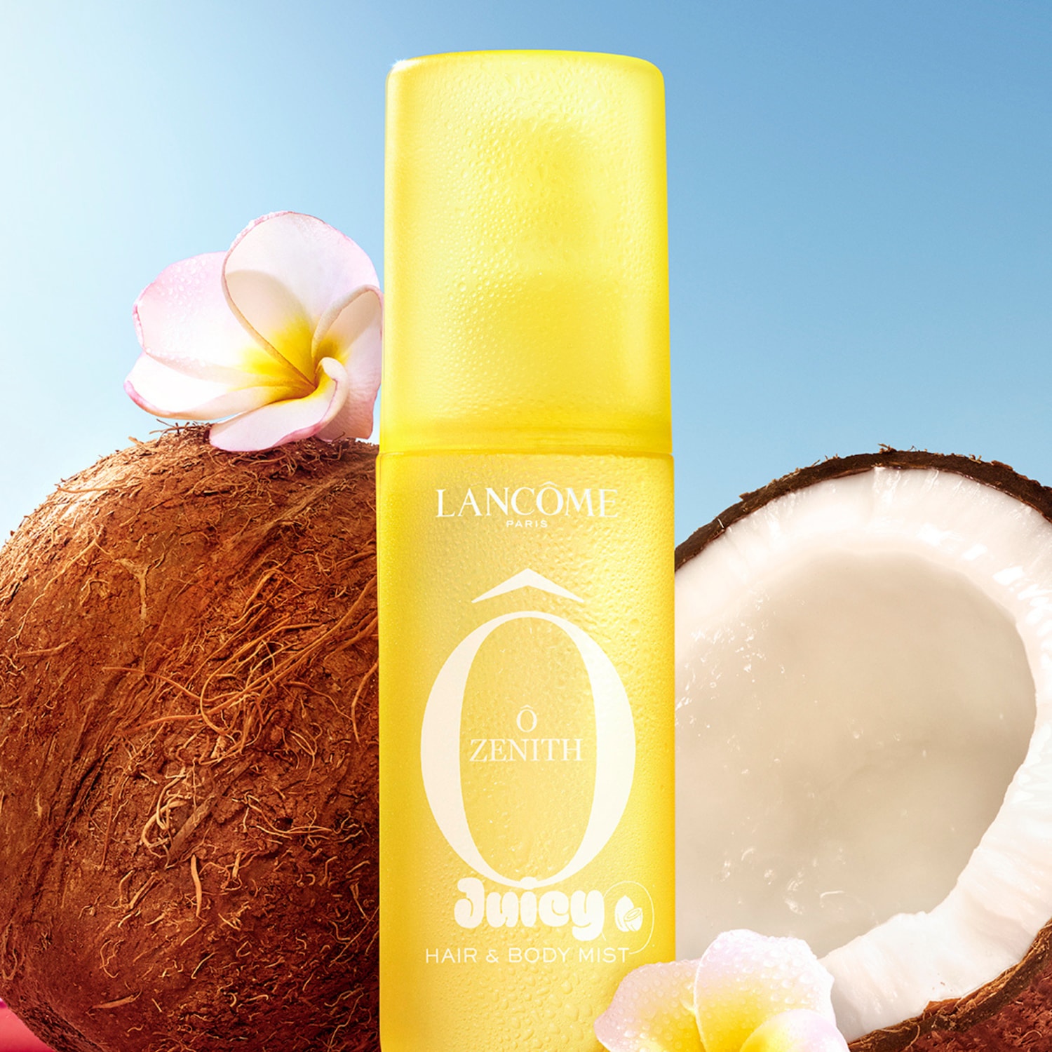 Le brume parfumée corps et cheveux Juicy & Zenith de Lancôme, dans un flacon jaune vif, est présentée avec des noix de coco et des fleurs de frangipanier, évoquant une ambiance tropicale.