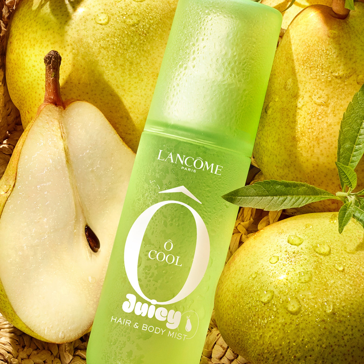 Le brume cheveux et corps Lancôme Ô Cool Juicy est présenté dans un flacon vert givré, orné de gouttes d'eau, et est entouré de poires fraîches et juteuses, dont une coupée en deux, sur un fond tressé.