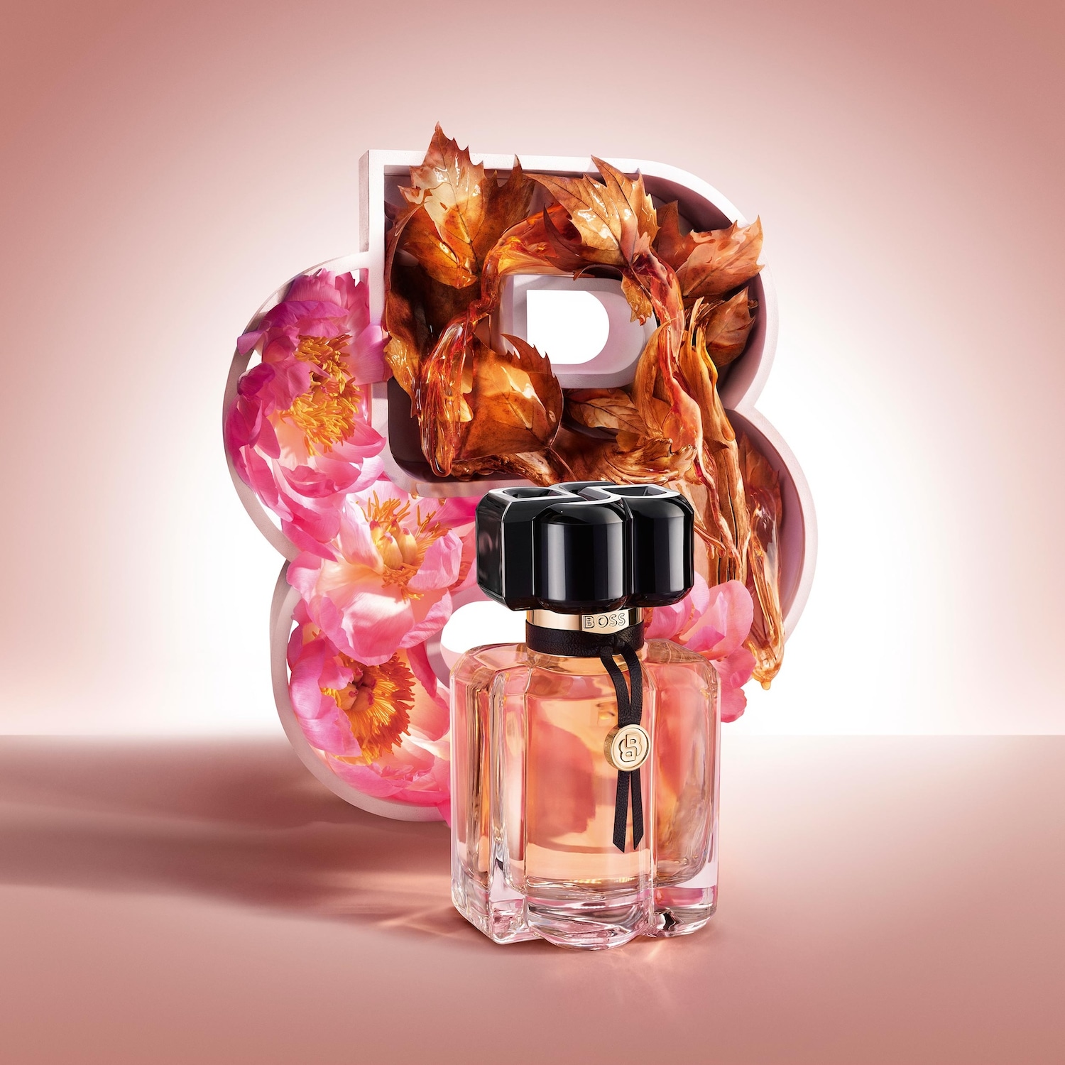 Ein BOSS The Scent Parfum Edition für Frauen, ein eleganter Flakon mit schwarzem Verschluss und einem goldenen B-Anhänger, steht vor einer großen weißen Acht, die mit rosa Blüten und braunen Blättern gefüllt ist.
