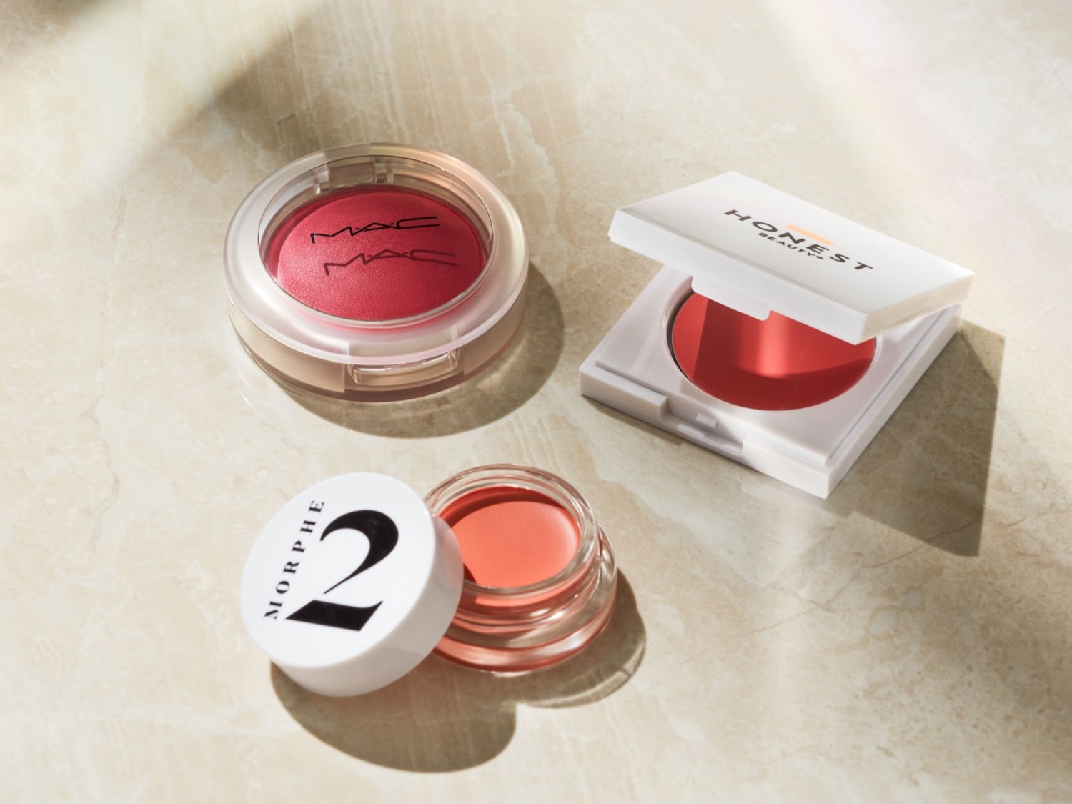 Drie blushes in verschillende tinten roze en perzik, waaronder een ronde MAC-blush in een transparant doosje, een vierkante Honest Beauty-blush in een wit doosje en een Morphe 2-crèmeblush in een glazen potje met een witte dop, liggen op een marmeren ondergrond.