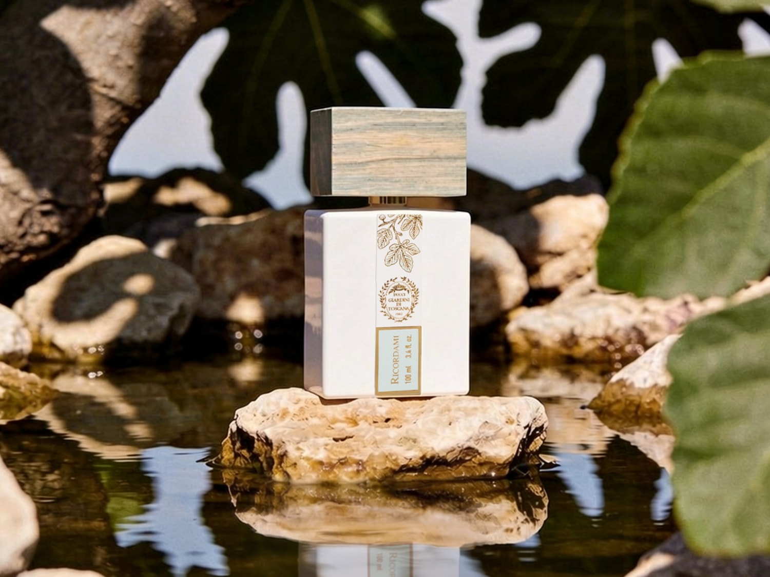 Een witte, rechthoekige parfumfles met een houten dop staat op een rots in het water, omringd door andere rotsen en groene bladeren, met de tekst "RICORDAMI 100 ml 3.4 fl. oz." en een gouden logo.