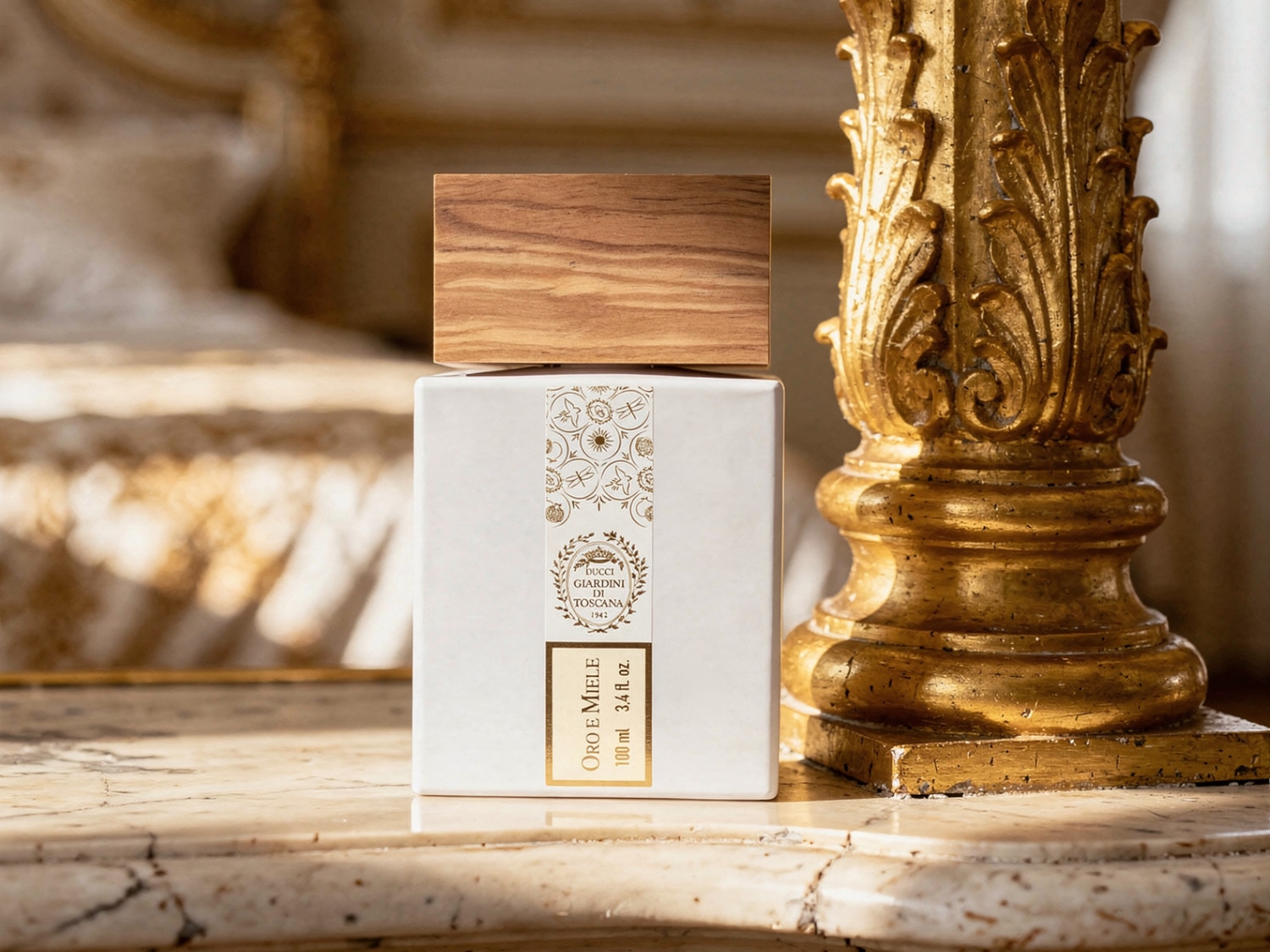 Een witte, vierkante parfumfles met een houten dop en een gouden etiket met de tekst "ORO E MIELE 100 ml 3.4 fl. oz." staat op een marmeren oppervlak naast een gouden, gedecoreerde pilaar.