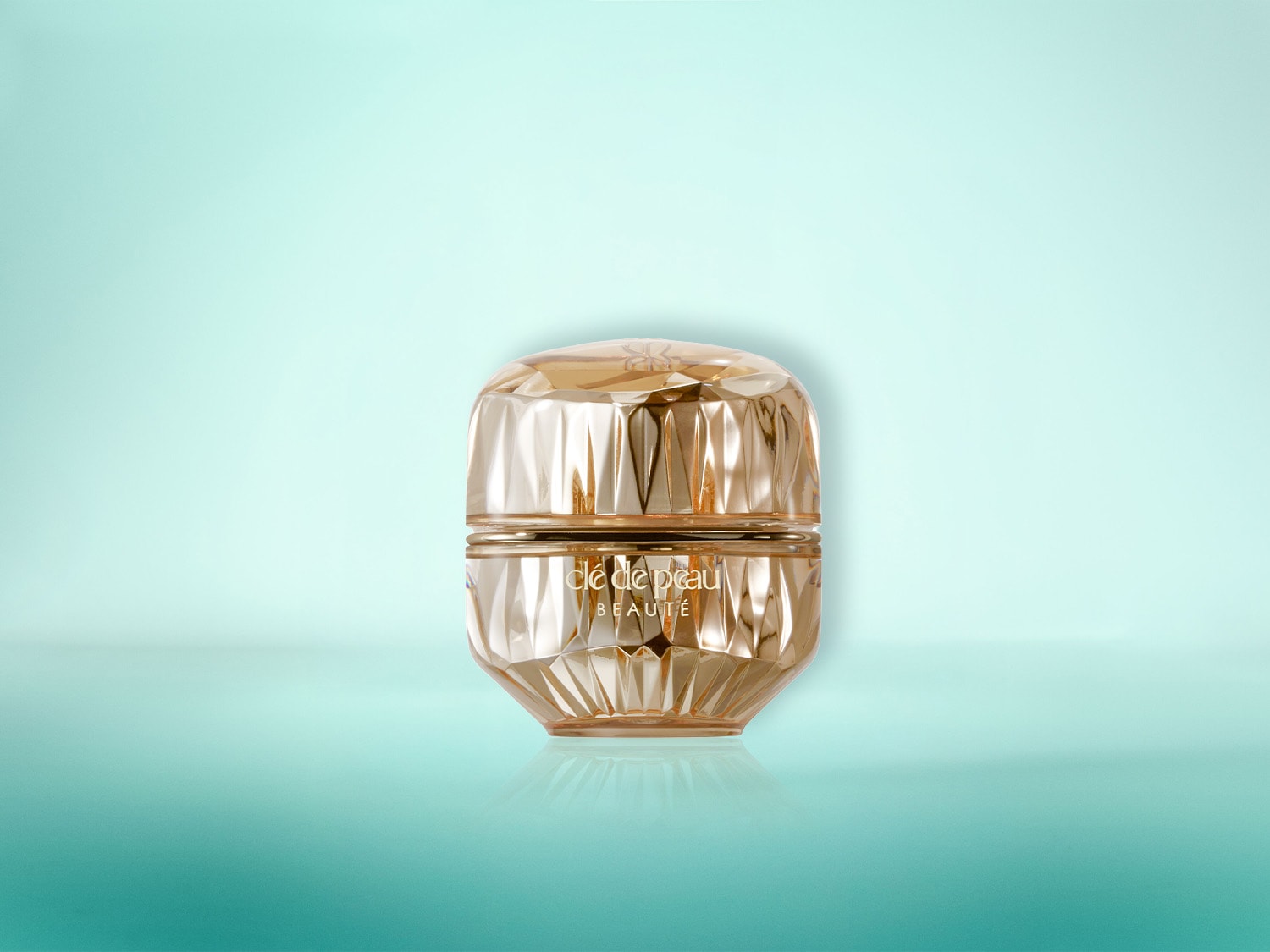 Ein luxuriöser goldener Tiegel von Clé de Peau Beauté mit facettiertem Design steht auf einer reflektierenden Oberfläche vor einem sanften blaugrünen Hintergrund.