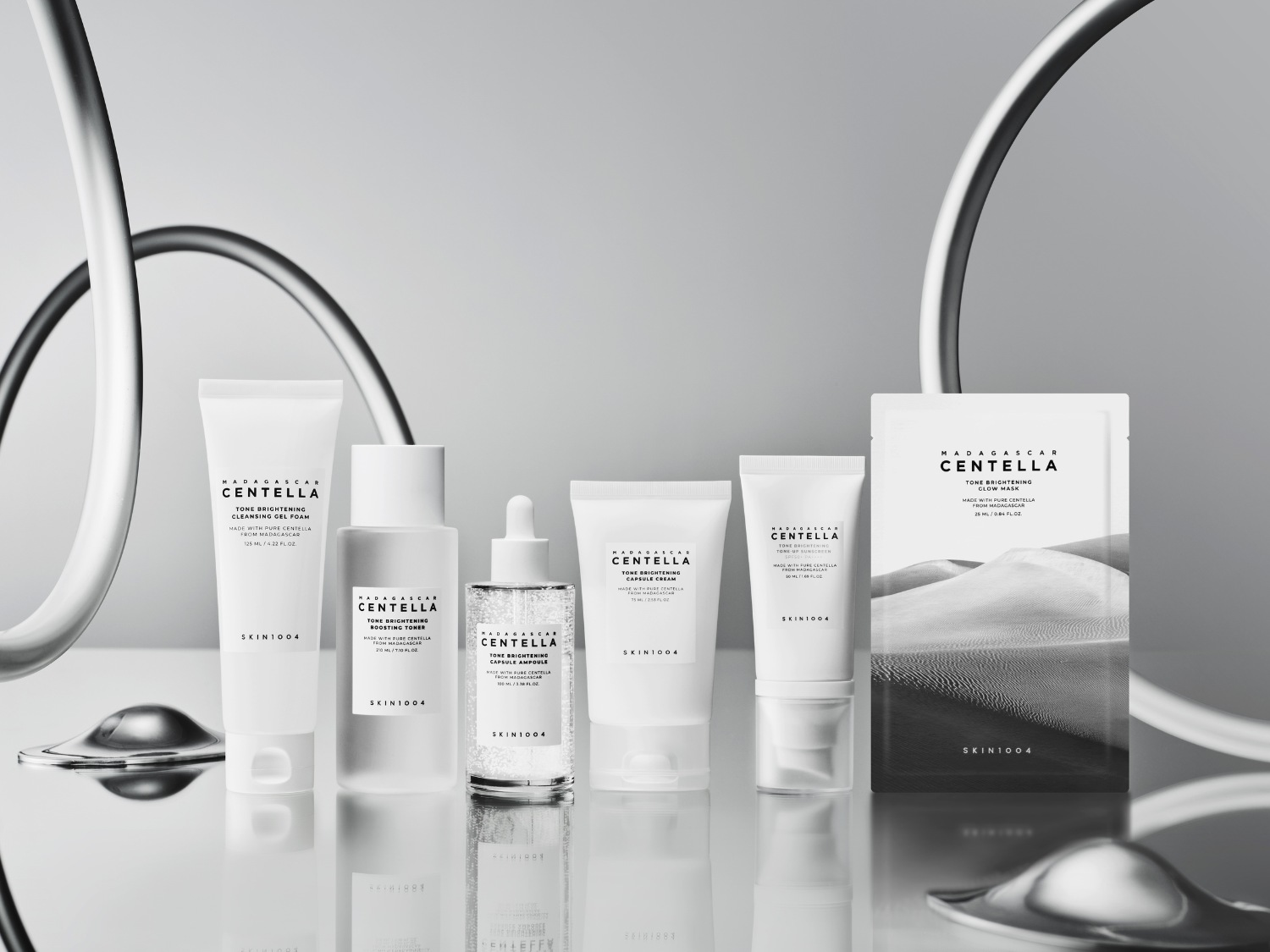 Kolekcja SKIN1004 Madagascar Centella Tone Brightening, zawierająca żel do mycia twarzy, tonik, ampułkę, krem i maskę w płachcie, jest prezentowana w eleganckich, białych opakowaniach z czarnymi napisami, ustawionych na błyszczącej powierzchni.