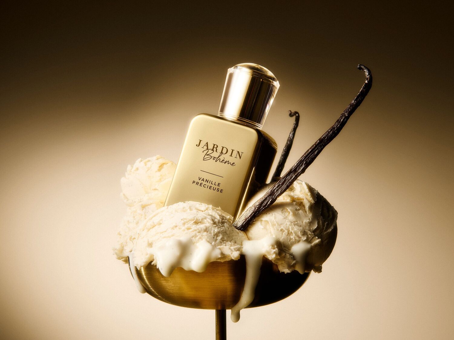 Le parfum Jardin Bohème Vanille Précieuse est présenté dans un flacon doré avec un bouchon assorti, posé sur des boules de glace à la vanille dans une coupe dorée, garni de gousses de vanille.
